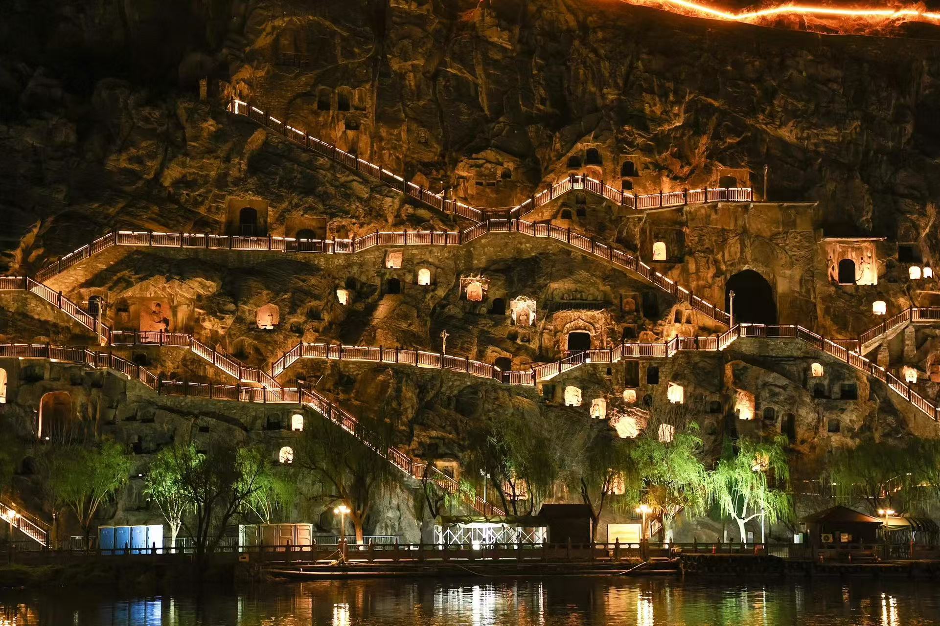 伊河對岸視角下的龍門石窟夜景:暖黃燈光點亮層層疊疊的窟龕,蜿蜒的棧道在崖壁間穿梭,燈光與水面倒影交相輝映,將千年石窟的靜謐與璀璨完美融合,氛圍感拉滿 伊河對岸視角下的龍門石窟夜景:暖黃燈光點亮層層疊疊的窟龕,蜿蜒的棧道在崖壁間穿梭,燈光與水面倒影交相輝映,將千年石窟的靜謐與璀璨完美融合,氛圍感拉滿