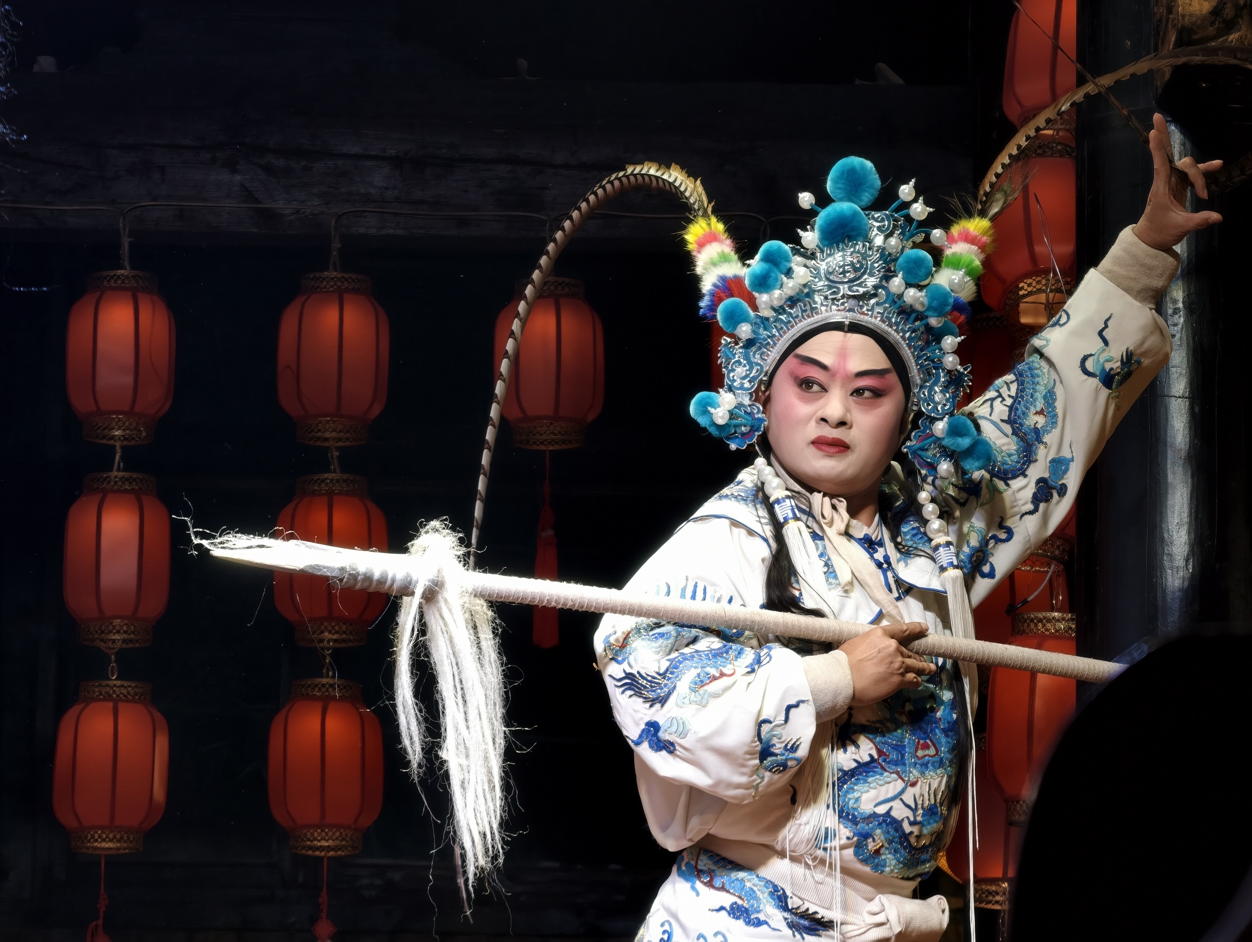 武生演員身著繡有藍龍的戲服，頭戴裝飾著絨球與翎子的盔頭，手持長槍，在紅燈籠的映襯下，擺出英武的造型，彷彿正演繹著一段沙場征戰的故事