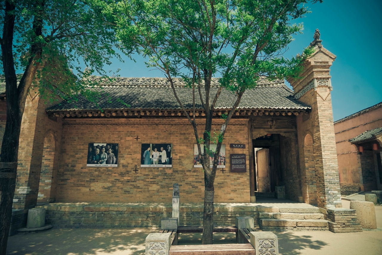 古舊的磚牆建築,牆上掛著劇照,搭配門前綠樹,還原關中民居場景,傳遞《白鹿原》影視文化底蘊,展現原著中的生活氛圍 古舊的磚牆建築,牆上掛著劇照,搭配門前綠樹,還原關中民居場景,傳遞《白鹿原》影視文化底蘊,展現原著中的生活氛圍