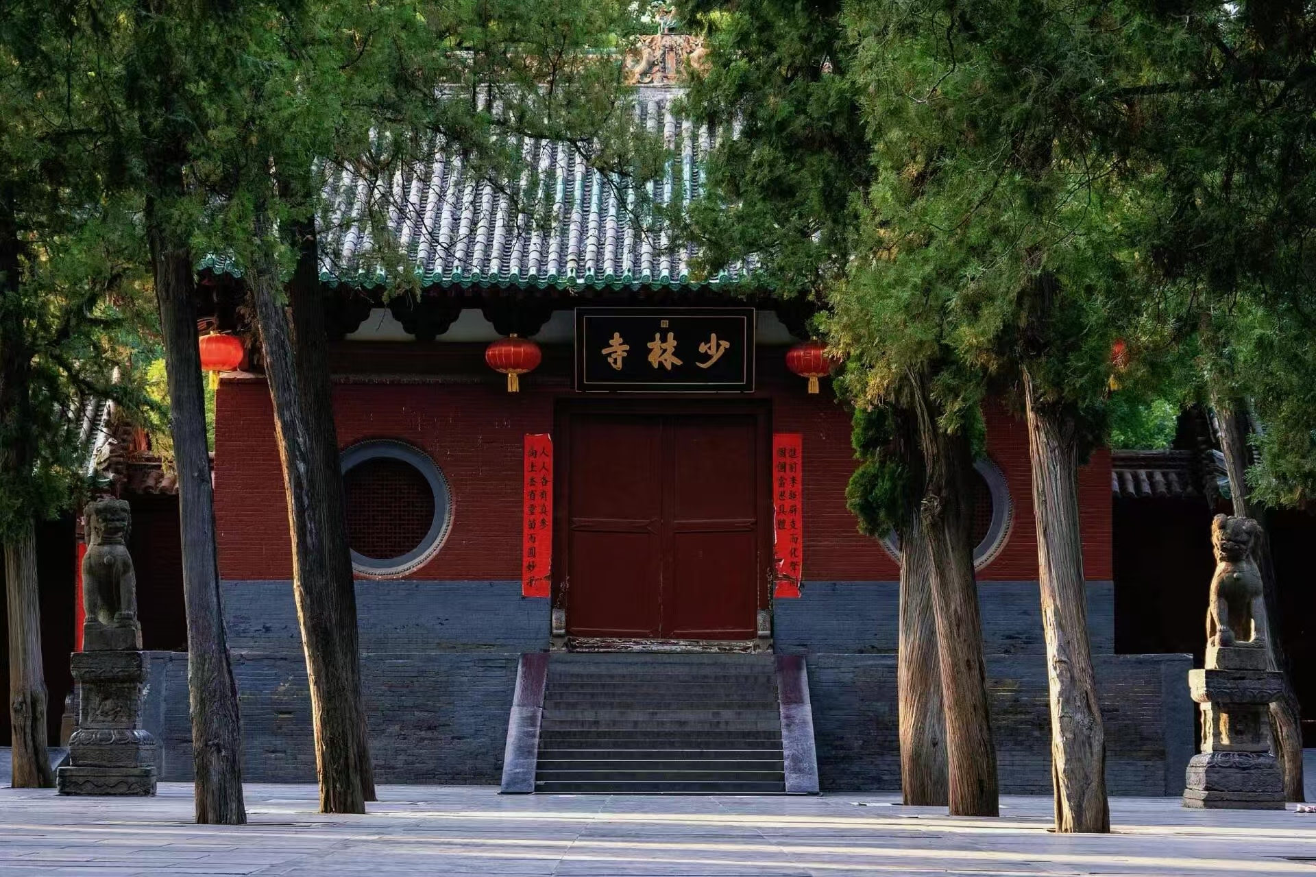 紅牆灰瓦的山門被蒼勁的古柏環繞，門楣上懸著 “少林寺” 黑匾，兩側掛著紅燈籠與紅底對聯，石獅子分立左右。陽光透過枝葉灑在地面，靜謐中透著禪意，是少林寺最具代表性的門面景緻