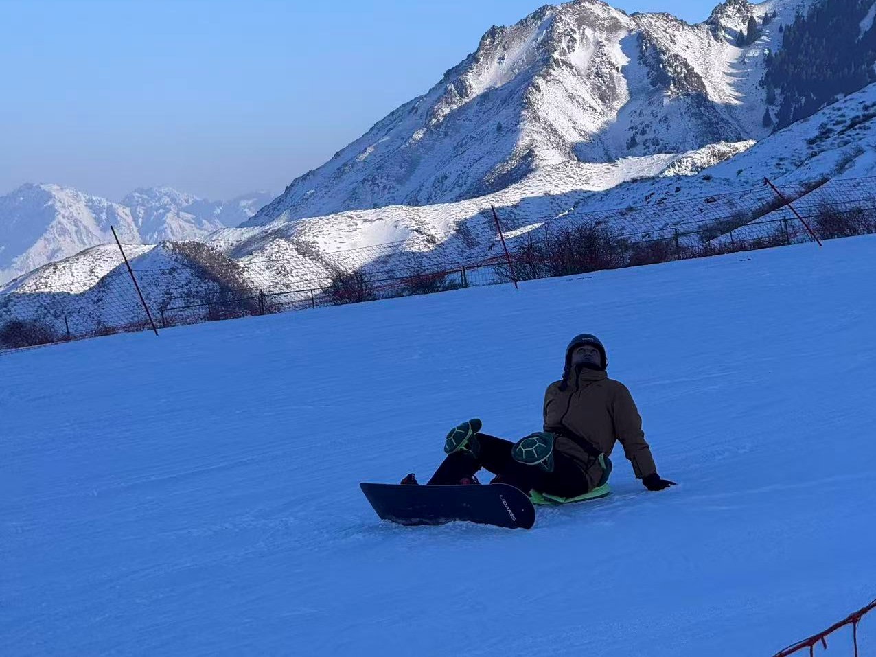 陽光斜照的雪坡上，雪友坐於雪地休憩，腳旁平放單板，佩戴烏龜造型護臀護膝。背景是巍峨錯落的覆雪山峰，防護網沿坡佈設，光影在山體勾勒明暗紋路，山野雪景壯闊，盡顯滑雪放鬆時刻