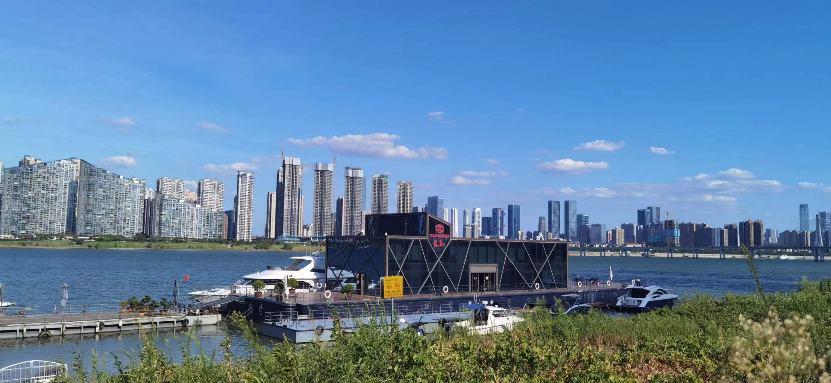On a clear day, the Xiangjiang River's surface is wide and expansive, and a modern-looking black floating dock building stands quietly in the heart of the river, with a white yacht peacefully docked at the berth.