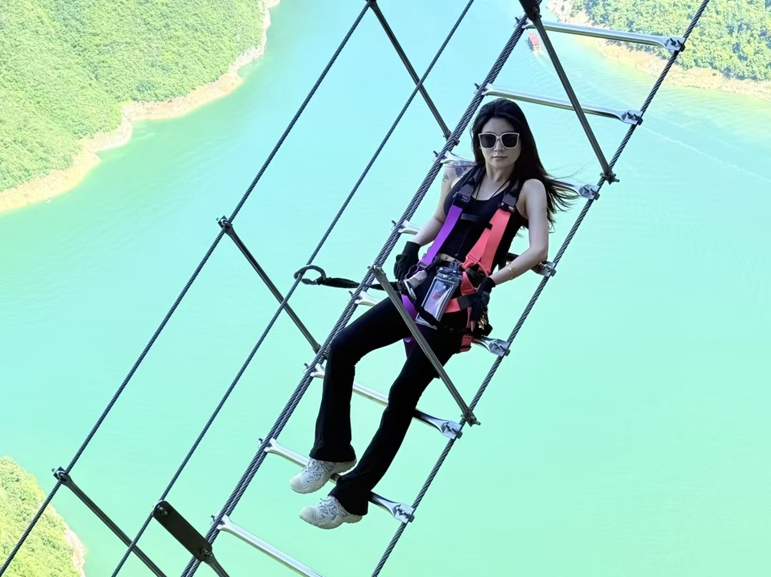 黑褲女子躺靠高空梯，長髮隨風揚，碧江在側，把高空險境變成了與山河私語的鬆弛時刻