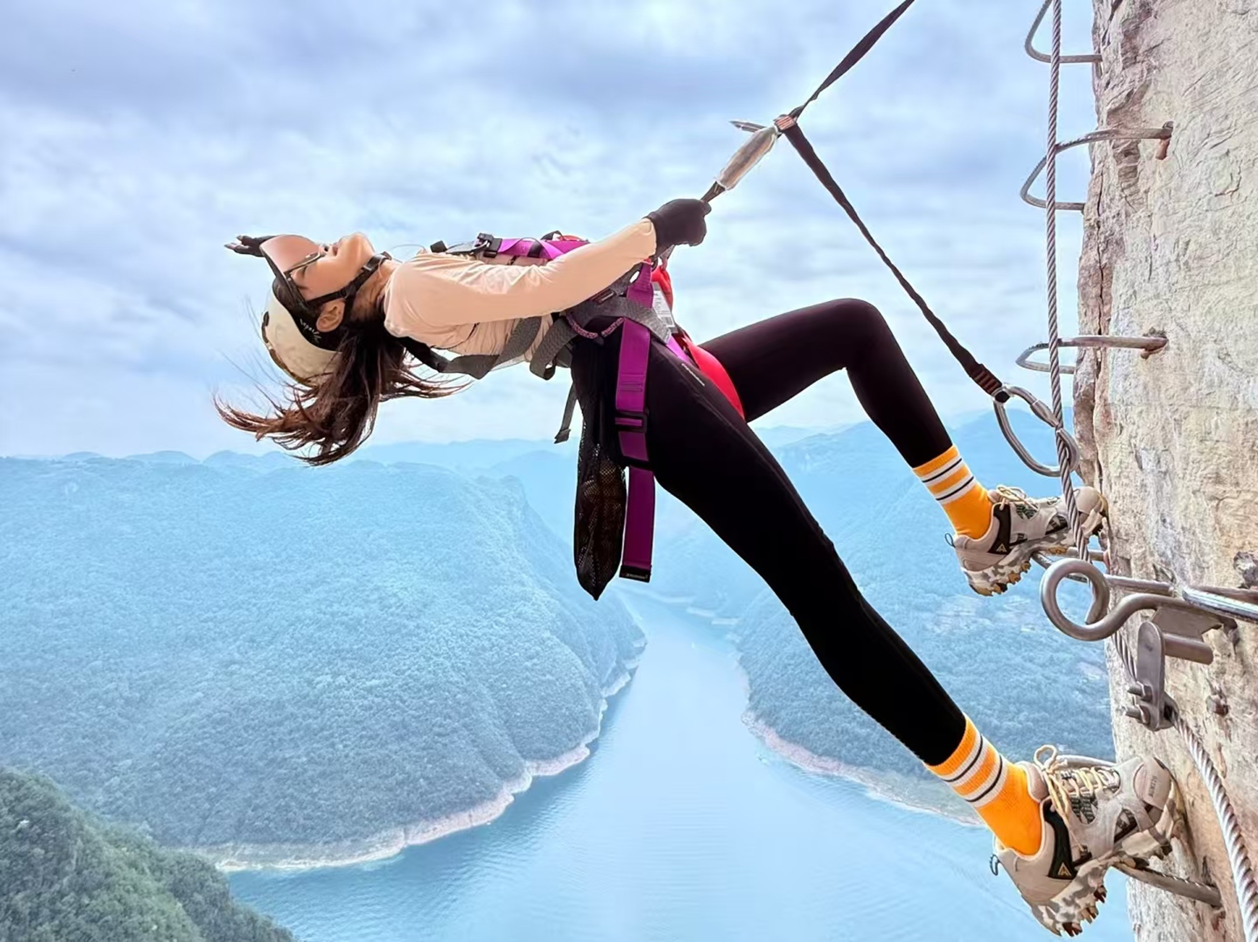 黃襪女子掛於崖壁梯，髮絲飛散，江灣隱於霧中，舒展姿態裹著懸崖挑戰的野性與詩意