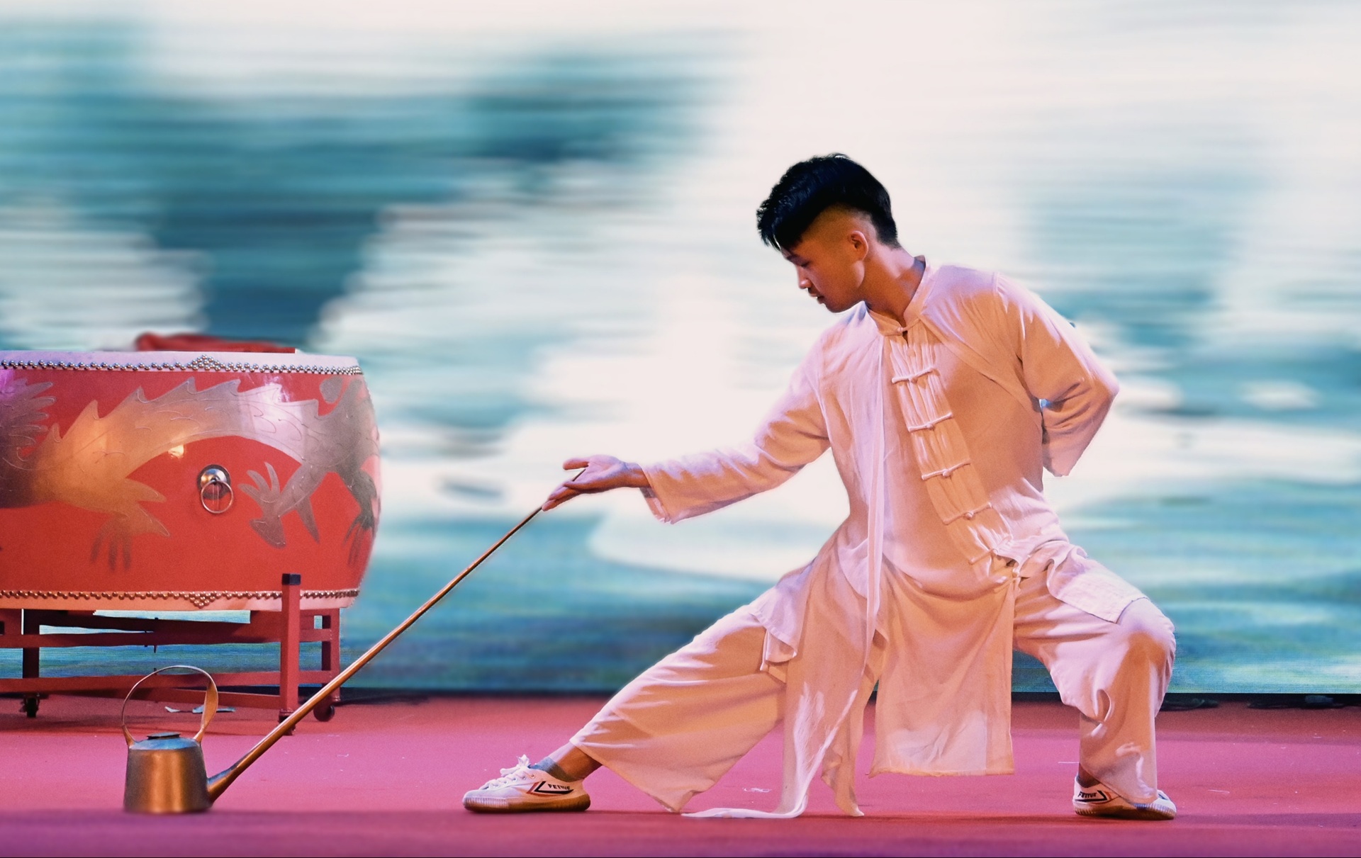 表演者穿中式白衣，單腿下蹲持長杆，旁置繪龍紅鼓，背景水色朦朧，動作舒展有韻味