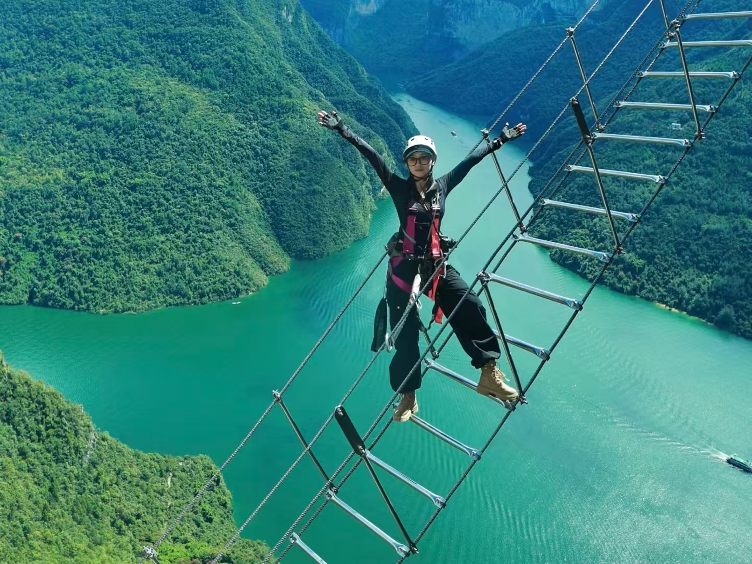 裝備齊全的人張開雙臂，立於峽谷高空的金屬梯上，青綠江面與翠嶺作背景，酷颯姿態裹著高空挑戰的刺激與壯闊