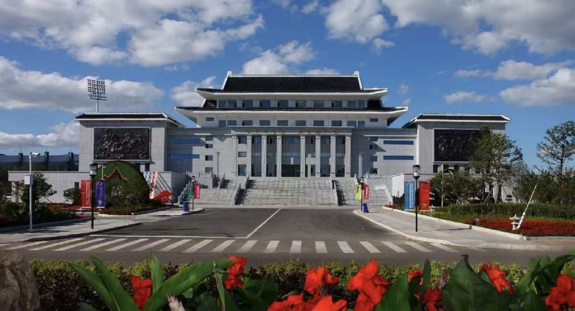 氣派的朝鮮族風格建築，灰白牆體搭配傳統飛簷屋頂，廣場開闊整潔。藍天白雲下，花壇鮮花盛放，兩側設有主題浮雕，是延吉具有代表性的文化地標建築