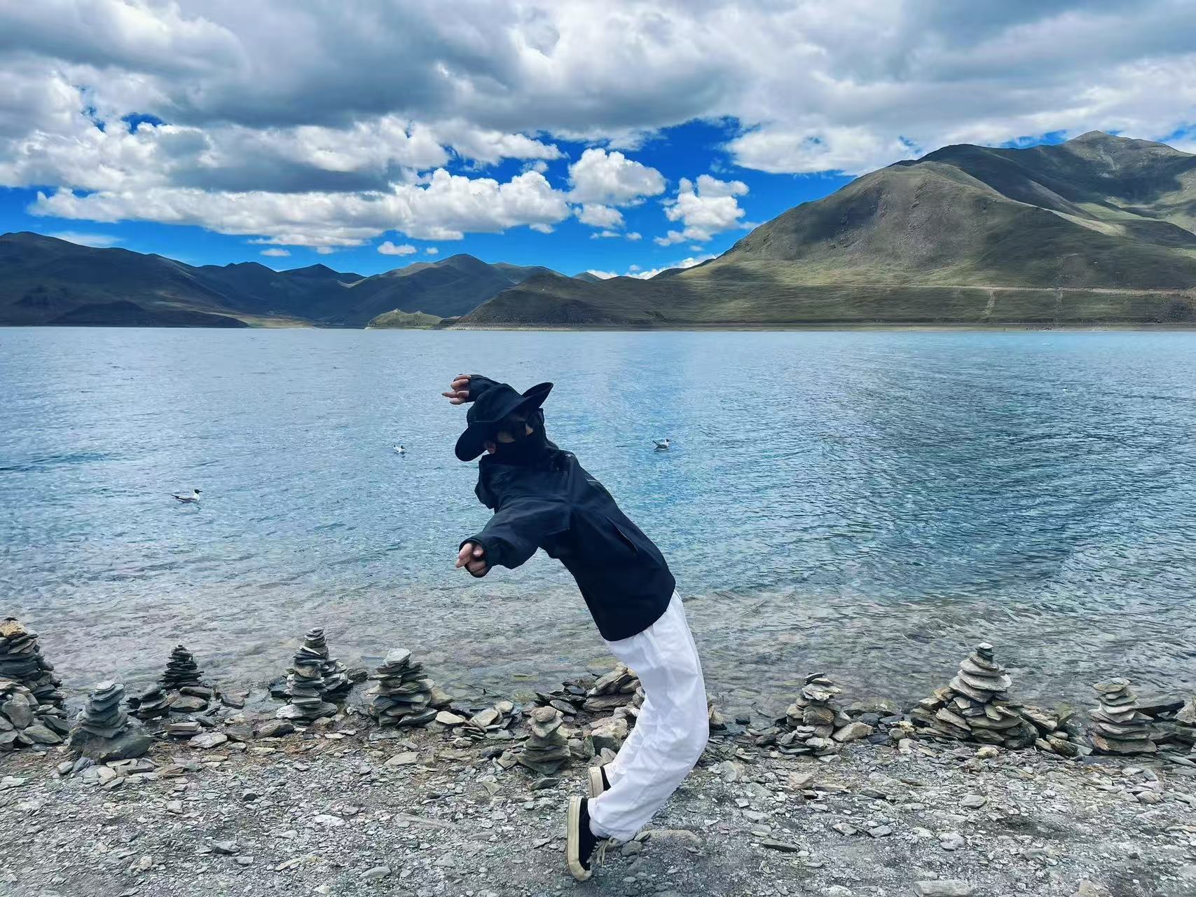 羊湖淺灘湖水清透透亮,岸邊堆疊祈福瑪尼堆。遊人黑衣白褲俏皮俯身擺拍,湖面水鳥自在浮游,遠處層疊青山襯著蓬鬆雲團,藍天碧水交融,滿是鬆弛的高原旅行氛圍感 羊湖淺灘湖水清透透亮,岸邊堆疊祈福瑪尼堆。遊人黑衣白褲俏皮俯身擺拍,湖面水鳥自在浮游,遠處層疊青山襯著蓬鬆雲團,藍天碧水交融,滿是鬆弛的高原旅行氛圍感
