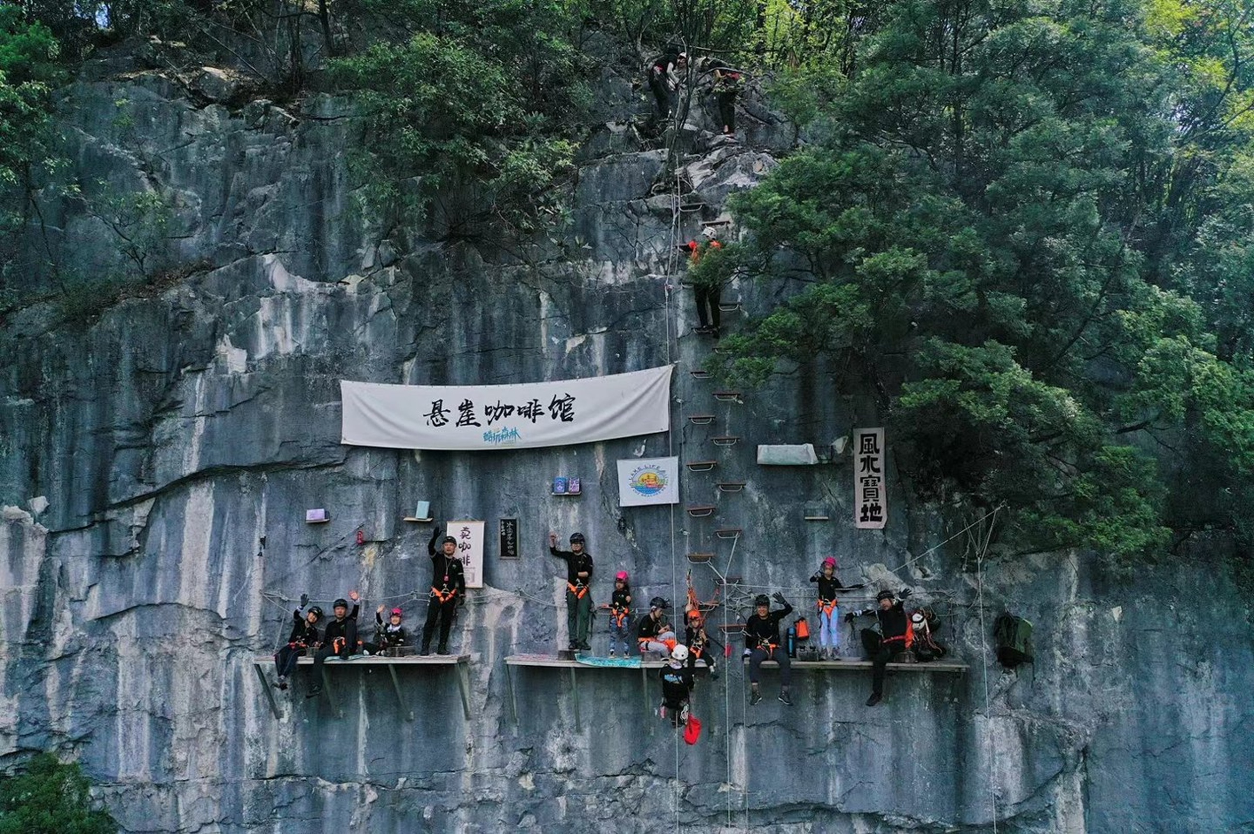 200 米高的懸崖咖啡館平臺上,遊客在巖壁前打卡,橫幅與攀巖繩相映,綠林作背景,是高空出片的絕佳機位 200 米高的懸崖咖啡館平臺上,遊客在巖壁前打卡,橫幅與攀巖繩相映,綠林作背景,是高空出片的絕佳機位