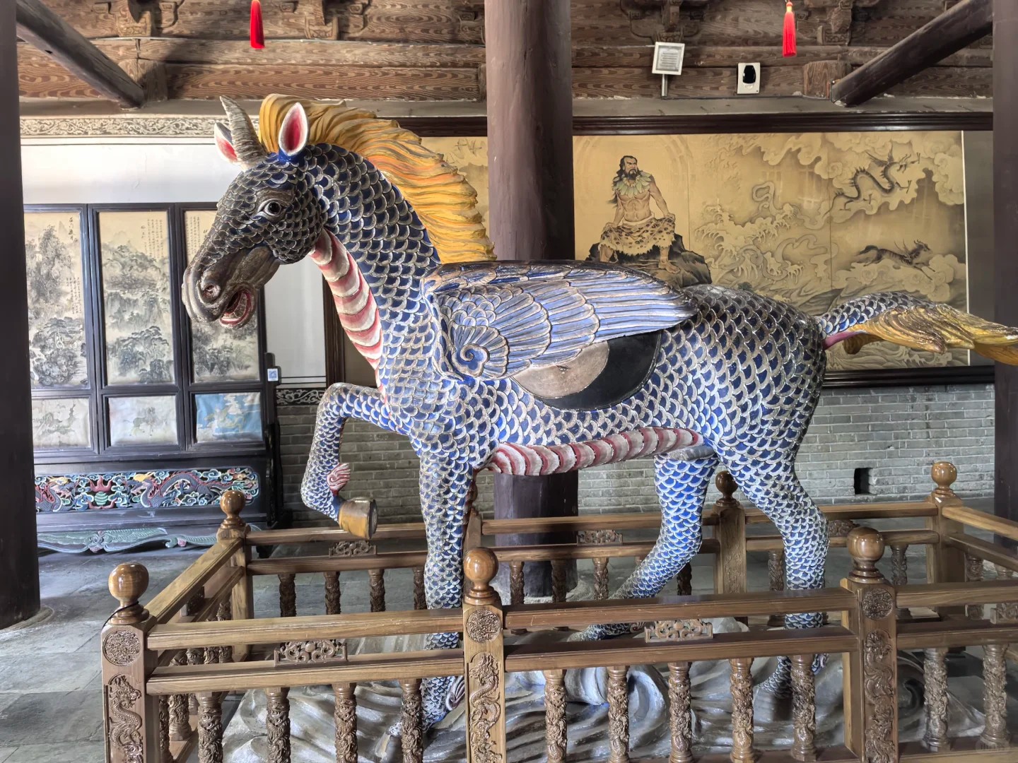 這是甘肅天水伏羲廟內的天馬雕塑景觀。畫面中央，一匹造型奇幻的“飛馬”騰躍於木質圍欄中，馬身覆蓋鱗片狀紋飾，背生羽翼，金黃鬃毛飛揚，融合了馬與飛鳥的意象，充滿神話色彩。雕塑所在建築為傳統明清風格，粗壯的樑柱、格窗與背景壁畫構成古樸的歷史空間，壁畫中隱約可見伏羲聖像，強化了人文始祖文化的氛圍。這尊雕塑以藝術化手法詮釋了“龍馬負圖出河”的伏羲傳說，將上古神話具象化，既展現了傳統雕刻工藝的精湛細節，也承載著中華文明對神獸圖騰的崇拜與想象，成為伏羲廟內獨特的文化符號。