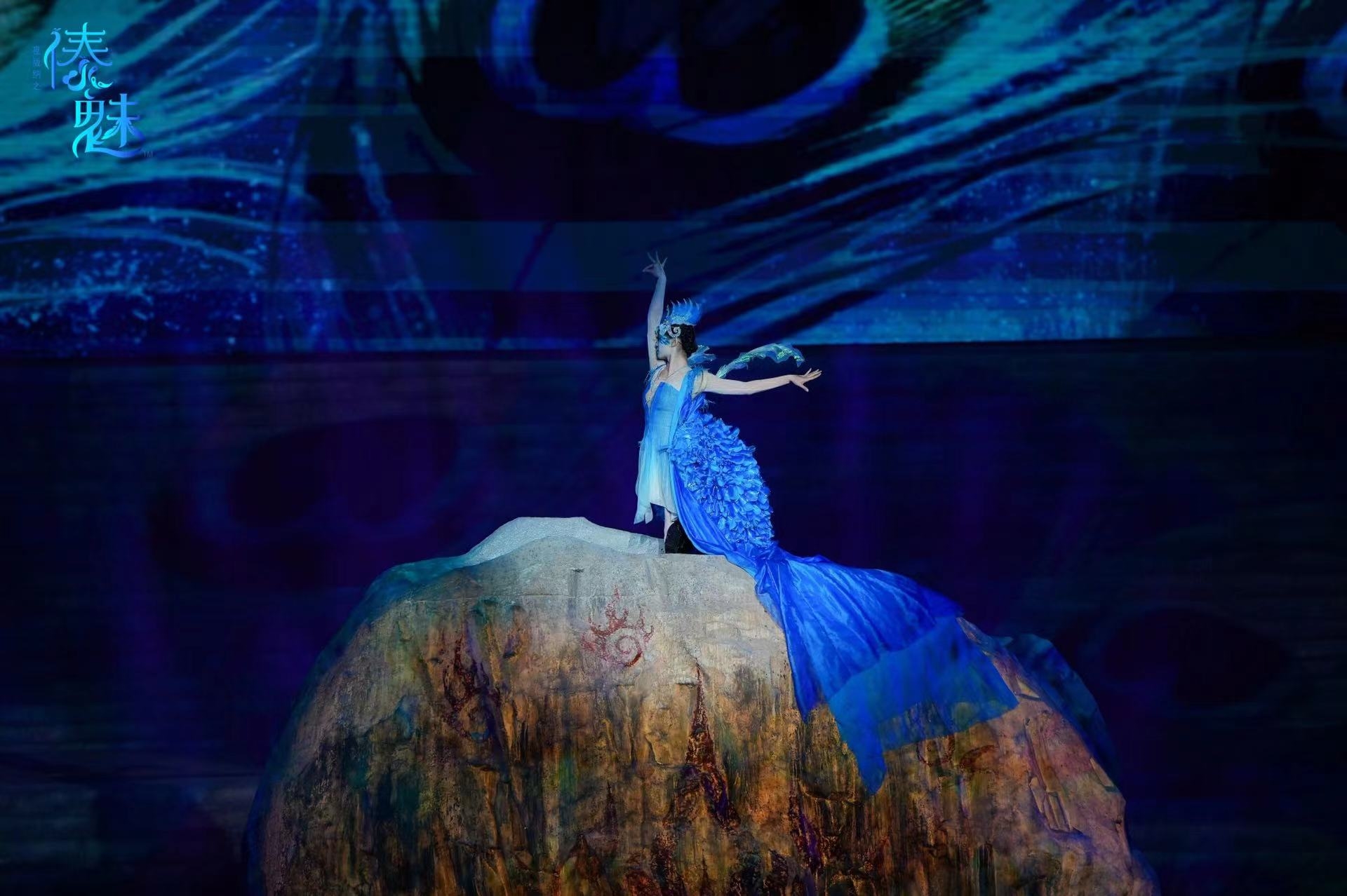 一位演員扮成孔雀造型,站在岩石狀道具上舞蹈,藍色的服飾如同孔雀羽毛,結合背景的光影效果 一位演員扮成孔雀造型,站在岩石狀道具上舞蹈,藍色的服飾如同孔雀羽毛,結合背景的光影效果