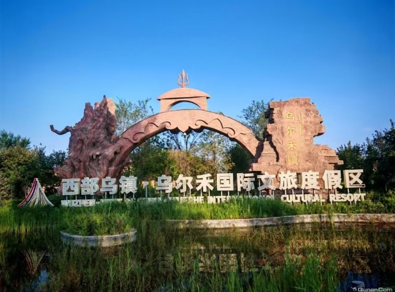 拱門造型充滿西域特色，搭配胡楊浮雕與烏爾禾鎮石刻，古樸大氣。門前水草繁茂，澄澈藍天映襯整片景觀。這裡毗鄰世界魔鬼城，是烏爾禾特色文旅小鎮入口，開啟北疆雅丹與小鎮休閒之旅
