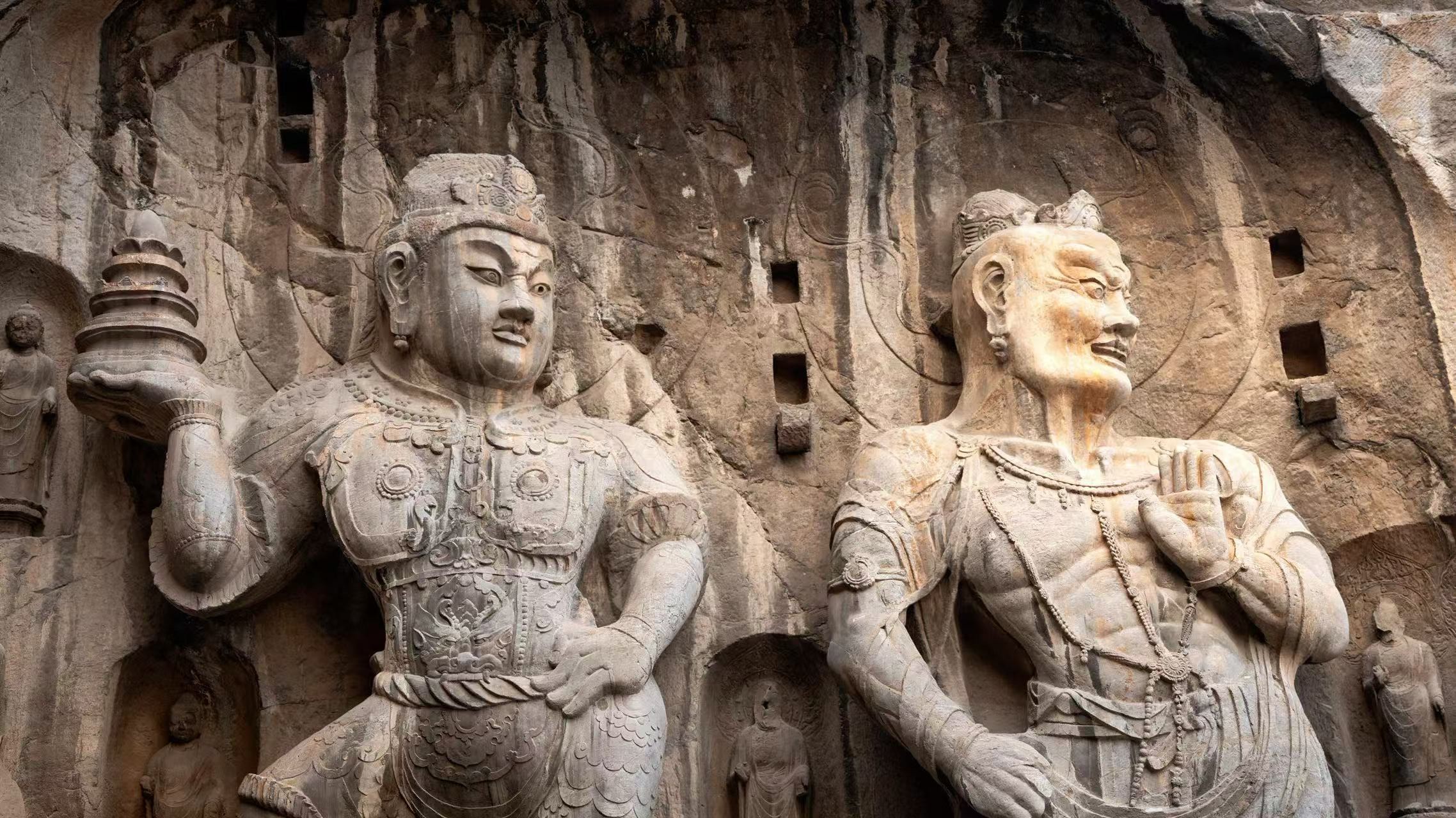 這是龍門石窟奉先寺的護法天王與力士像特寫:左側天王身披鎧甲、手託佛塔,神情威嚴;右側力士袒胸露腹、肌肉虯結,怒目圓睜。二者均依山而鑿,石刻紋理與風化痕跡盡顯千年厚重,是唐代造像剛勁與力量感的典型代表 這是龍門石窟奉先寺的護法天王與力士像特寫:左側天王身披鎧甲、手託佛塔,神情威嚴;右側力士袒胸露腹、肌肉虯結,怒目圓睜。二者均依山而鑿,石刻紋理與風化痕跡盡顯千年厚重,是唐代造像剛勁與力量感的典型代表