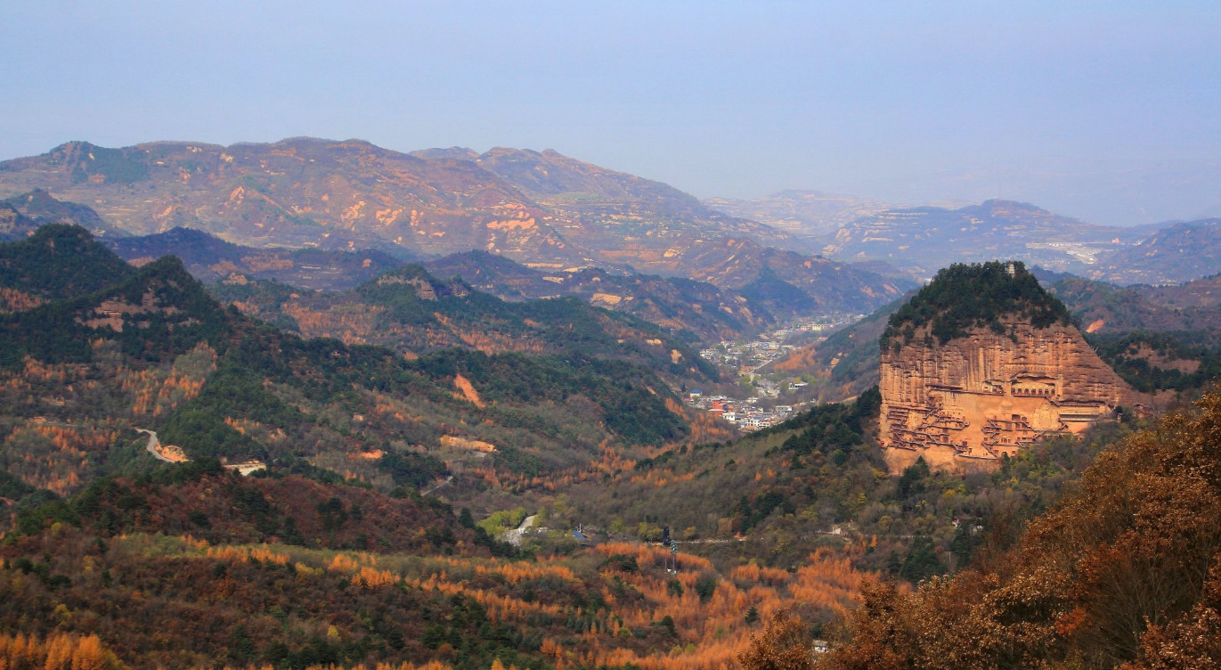 麥積山孤峰突兀於層巒疊嶂之間，山體呈赭紅色，與四周漸染秋色的山林形成鮮明對比，橙黃交織的植被為群山披上暖色華服。山體表面隱約可見密集的洞窟遺蹟，見證著千年佛教藝術的沉澱。遠眺山腳，城鎮隱於山谷，自然與人文和諧共生。作為絲綢之路上的世界文化遺產，麥積山以獨特的丹霞地貌為基底，將自然奇觀與石窟文化完美融合，既呈現了隴原秋日的絢爛畫卷，也鐫刻著跨越十六國的歷史與藝術輝煌。