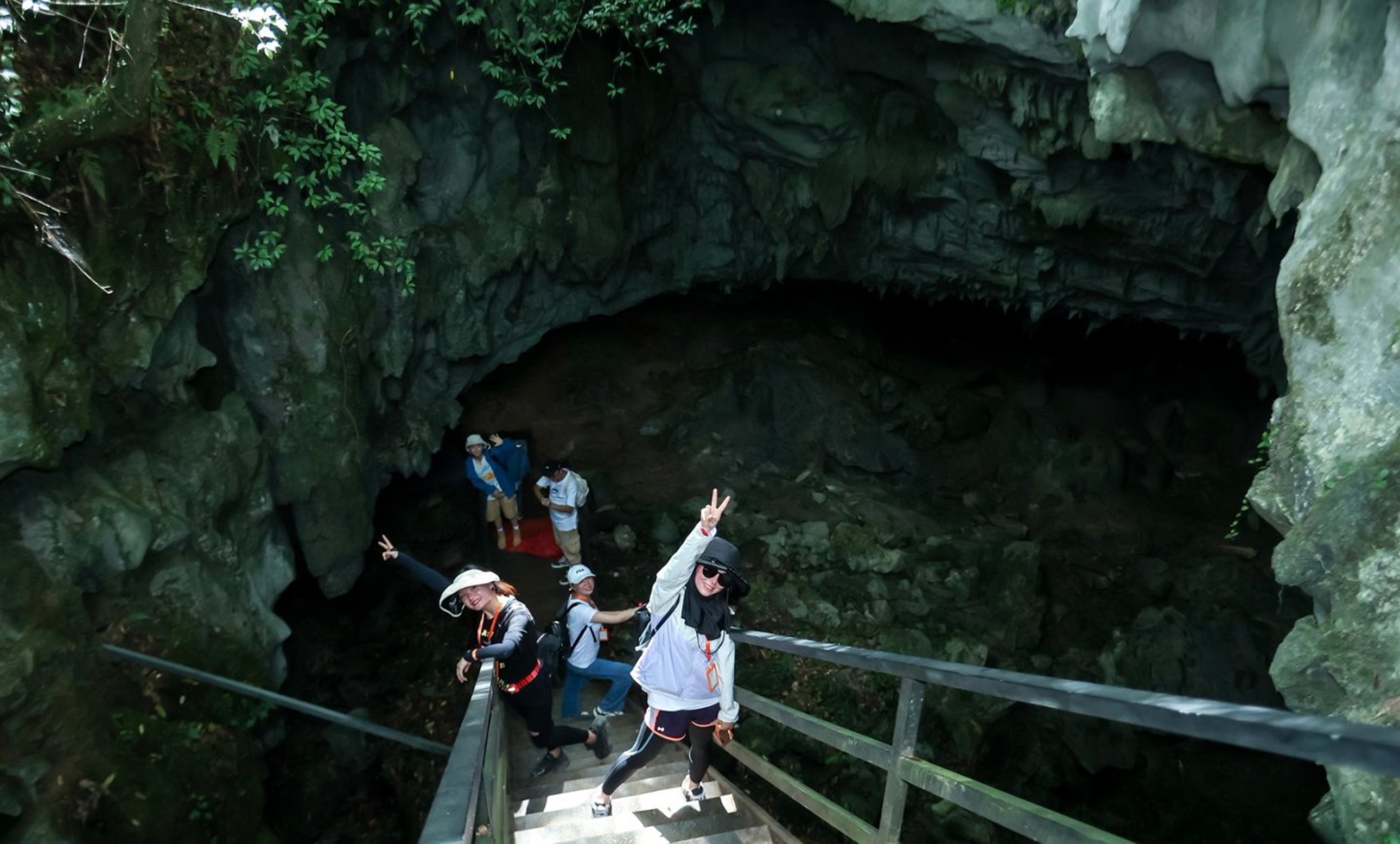 遊客沿階梯步入溶洞洞口,有人興奮比耶,巖壁綠植與洞穴暗影相映,是踏入地心探險前的期待與雀躍時刻 遊客沿階梯步入溶洞洞口,有人興奮比耶,巖壁綠植與洞穴暗影相映,是踏入地心探險前的期待與雀躍時刻
