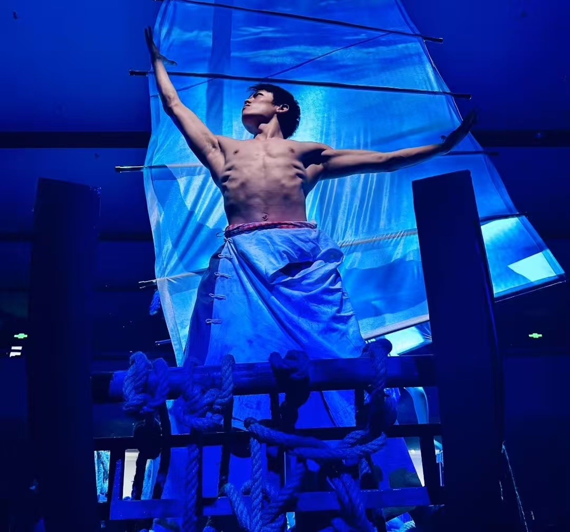 演員赤裸上身著藍布裙，立於船型道具上展臂，藍調燈光襯出漁家舞臺氛圍，張力十足
