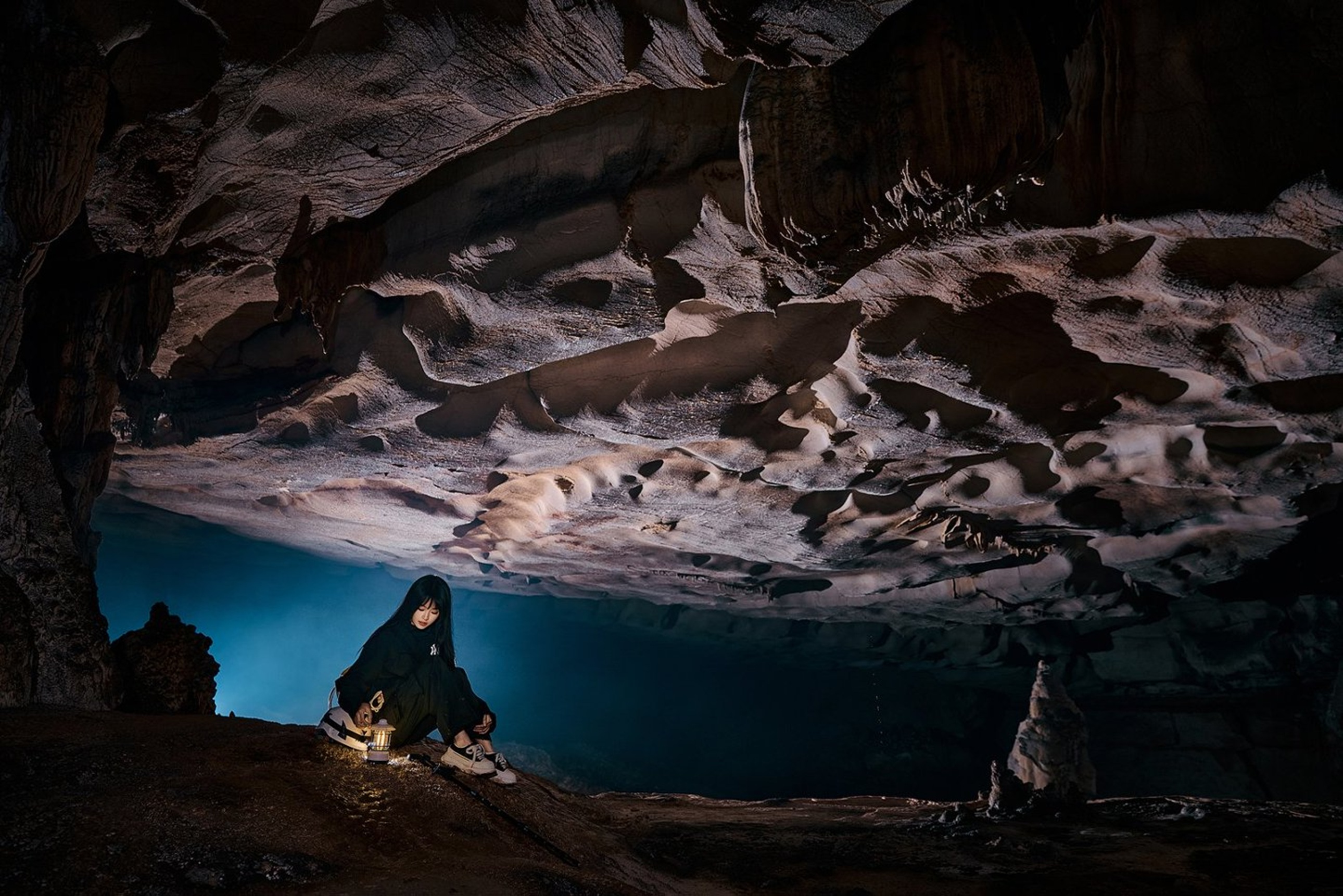 女孩蹲在溶洞深處,手捧煤油燈,巖壁如層疊雲朵般舒展,藍調背景與暖黃燈光交織,盡顯喀斯特地貌的奇幻與靜謐 女孩蹲在溶洞深處,手捧煤油燈,巖壁如層疊雲朵般舒展,藍調背景與暖黃燈光交織,盡顯喀斯特地貌的奇幻與靜謐