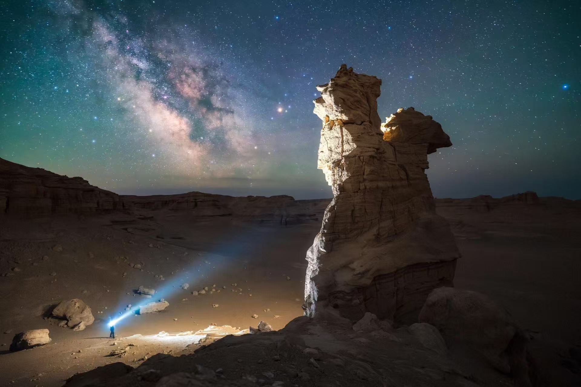 深邃夜空，璀璨銀河橫跨天際。一塊造型獨特的風蝕奇石矗立荒漠，底部有人手持手電打出光束。星光與地燈交織，孤寂雅丹，營造出星際秘境的浪漫氛圍感