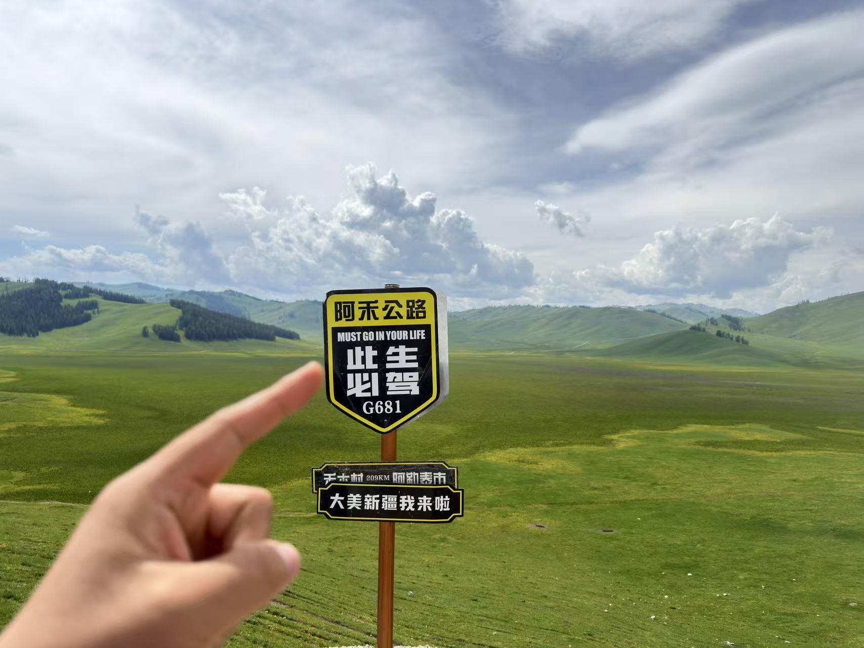 畫面中心立著 G681 阿禾公路網紅標牌，標註此生必駕與兩地裡程。一隻手指向路牌，背後是無邊青綠草原與層疊青山，白雲鋪滿藍天，是自駕途中標誌性打卡點位