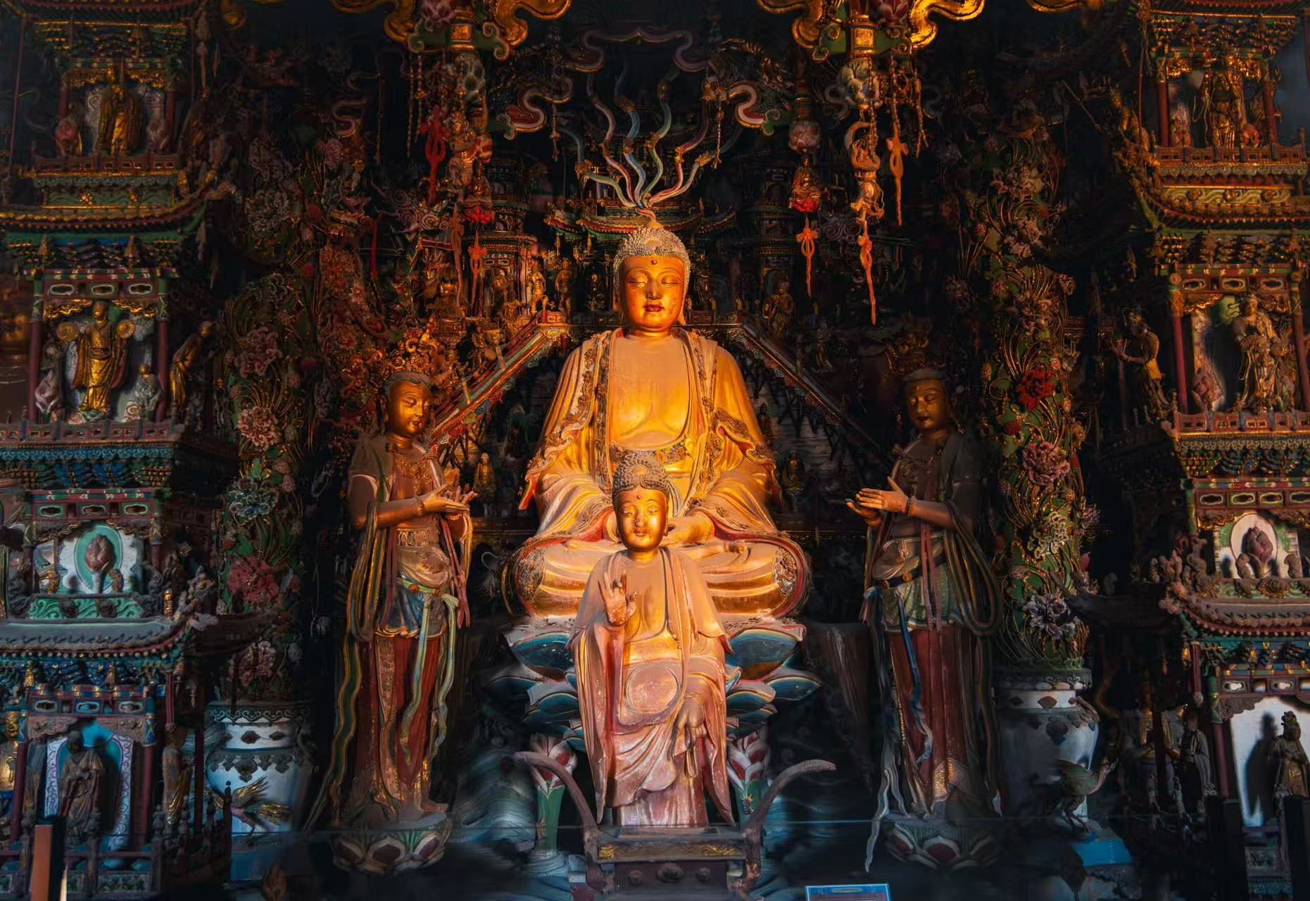 小西天殿堂正中端坐鎏金主佛，面容溫潤祥和，身前立小型佛像，兩側侍立彩繪菩薩，身後層層懸塑樓閣、仙佛環繞，光影明暗交織，營造出靜謐莊嚴的佛國氛圍
