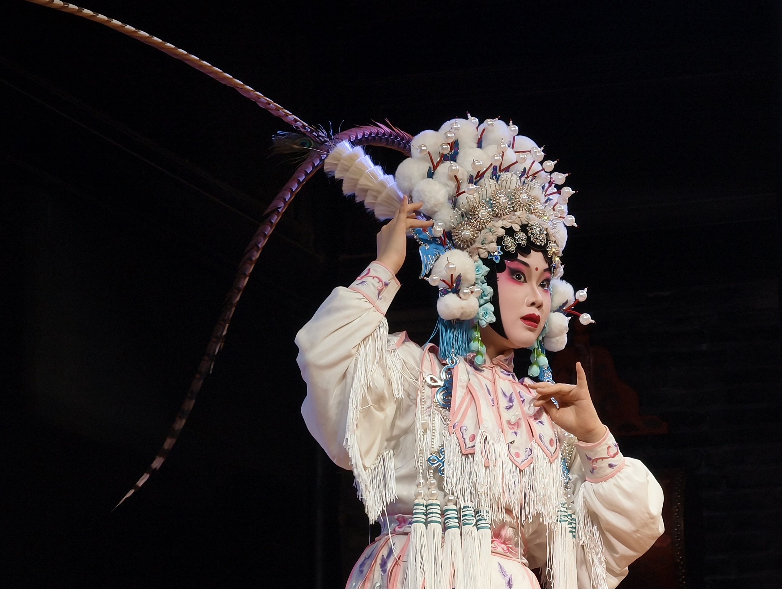 旦角演員頭戴精緻華麗的頭飾，綴滿絨球與珠翠，長長的翎子隨風微動，一身繡滿花紋的戲服盡顯典雅，她正以優美的姿態演繹著戲曲中的經典角色，眉眼間的妝容與動作皆充滿戲曲韻味