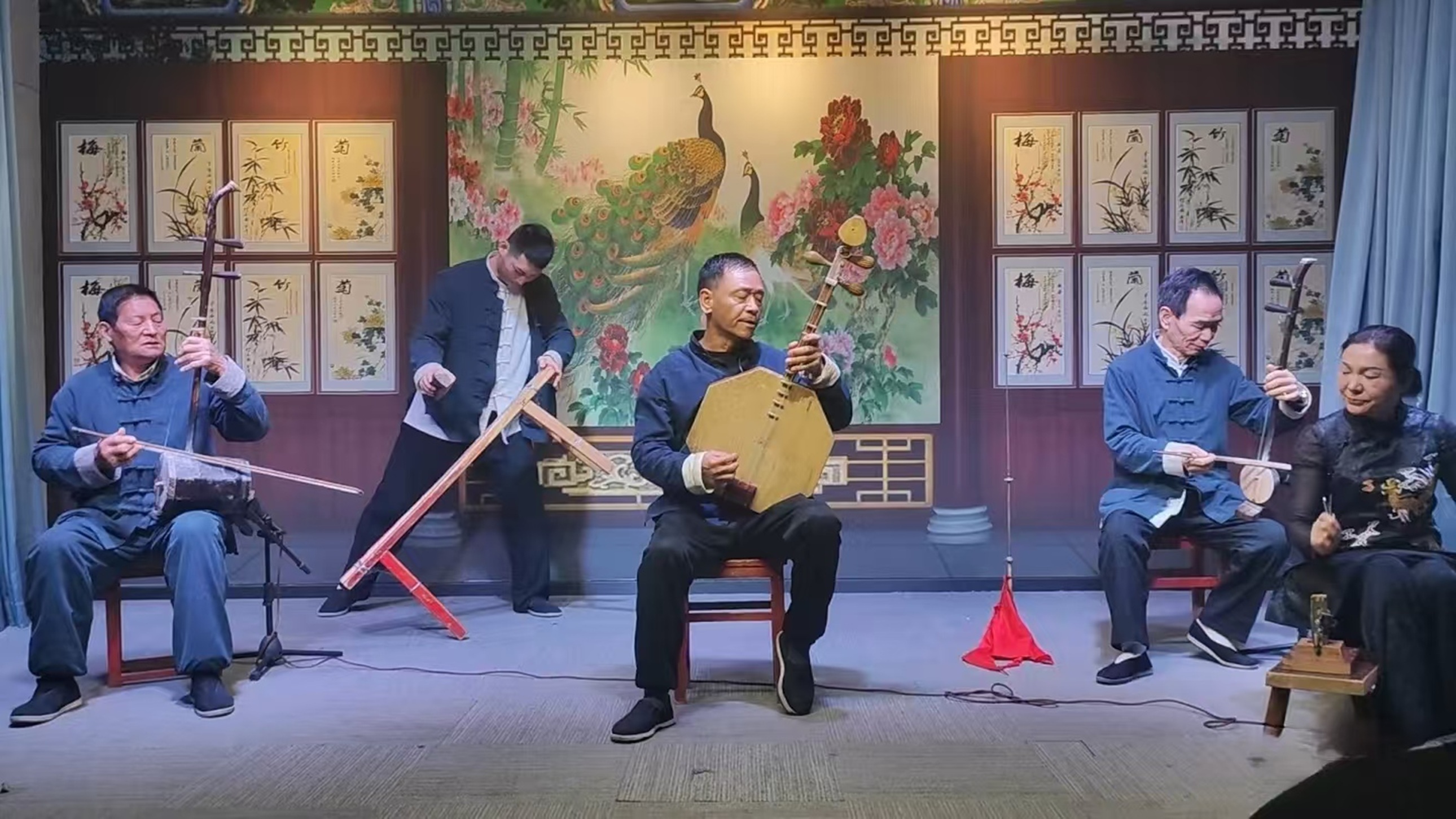 五位樂師著中式服飾，操持板胡、三絃等傳統樂器，在梅蘭竹菊裝飾的背景前演奏，盡顯戲曲伴奏的韻味