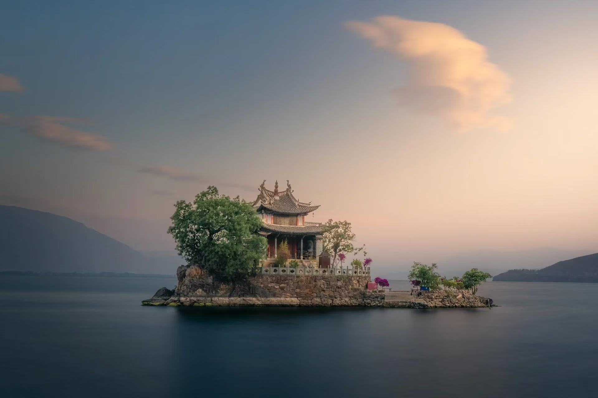 洱海上的小普陀孤懸湖面，中式古建立於礁石小島，古樹相伴。柔和晨昏天光漫過天際，遠山淡淡隱於霧色，平靜湖水託舉小島，古樸靜謐，自帶空靈悠遠的東方詩意