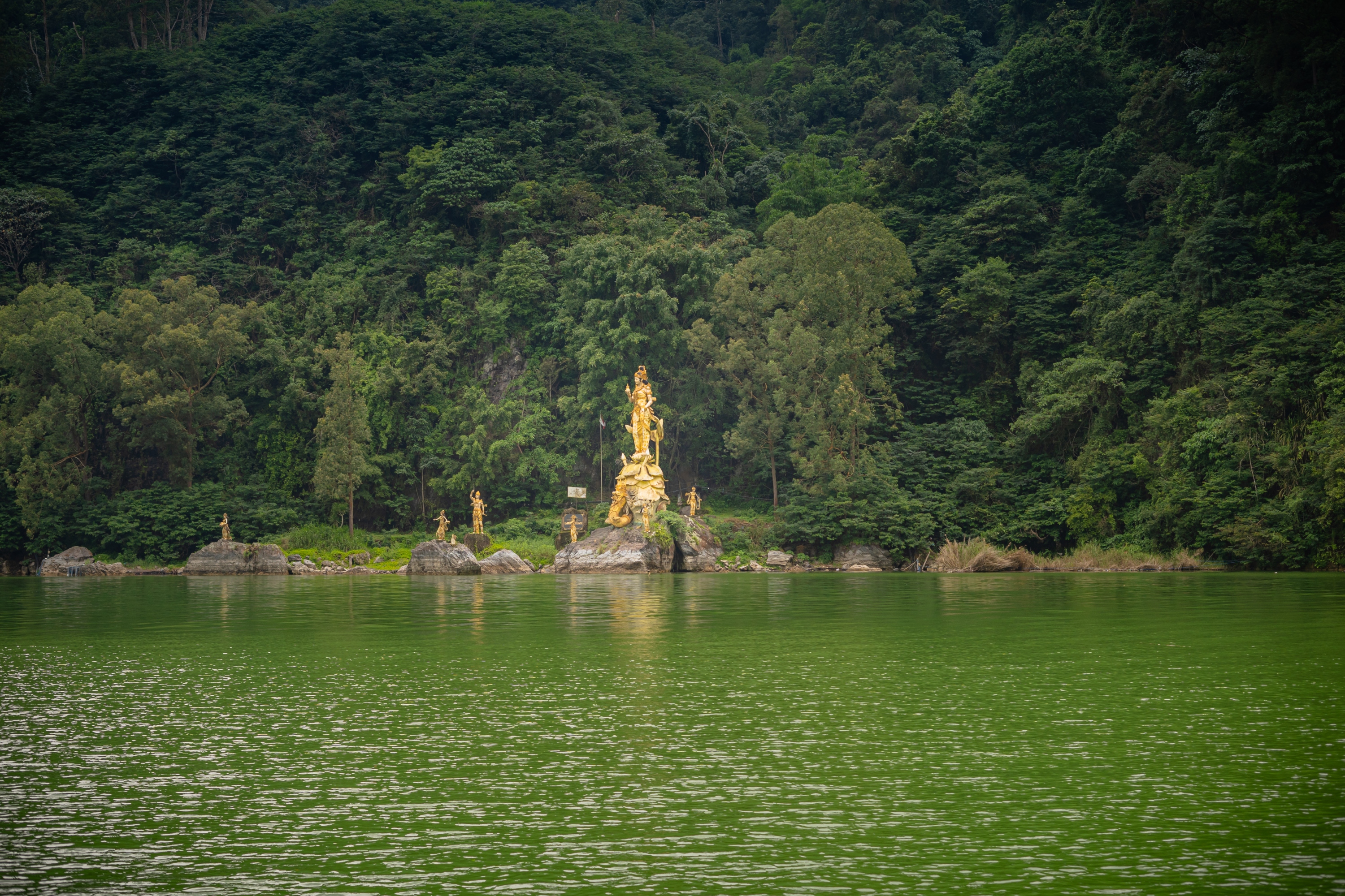 金色的雕像雄偉地矗立在湖岸邊，周圍環繞著茂密的森林，更增添了莊嚴肅穆的氛圍。