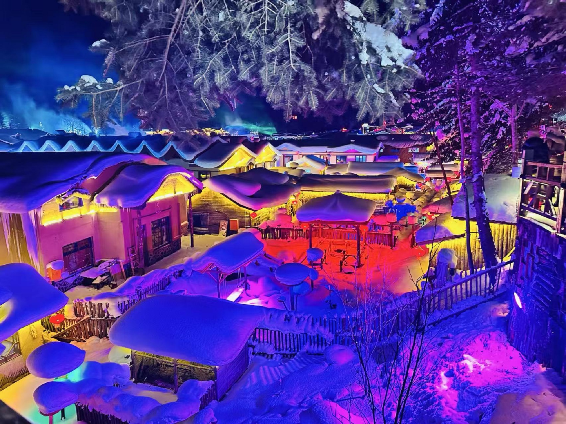 夜幕籠罩雪鄉，木屋屋頂堆積厚實蘑菇雪，五彩燈光灑落院落。木柵欄、涼亭被皚皚白雪包裹，松樹覆滿積雪，絢爛夜色勾勒出夢幻冰雪農家院落，氛圍感十足