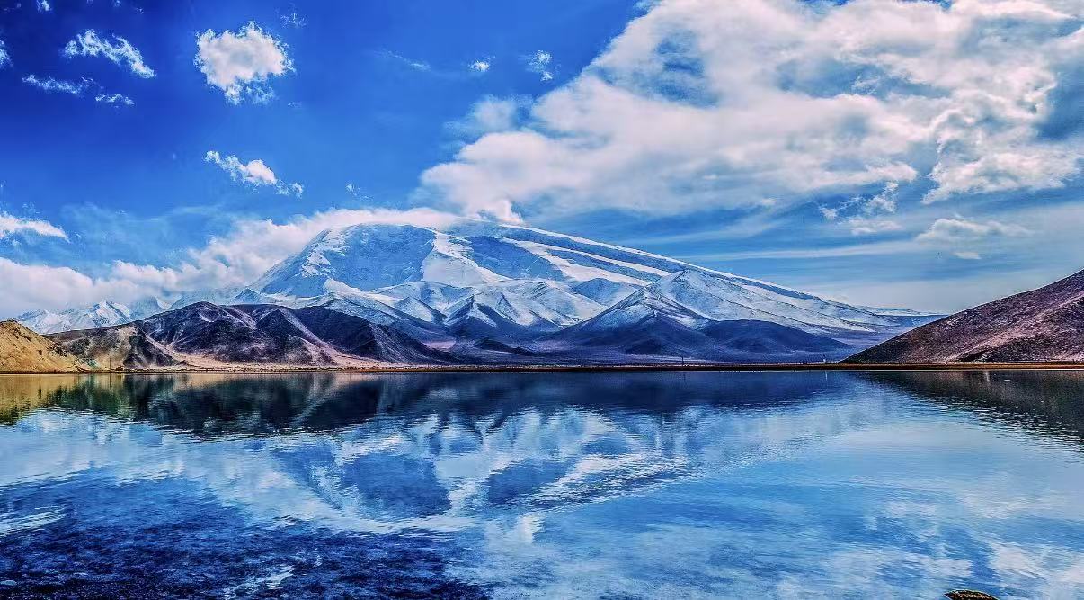 慕士塔格峰巍峨矗立，皚皚雪峰倒映在喀拉庫勒湖如鏡的水面上，藍白相映，純淨又震撼。遠處氈房點綴湖畔，天空流雲輕舞，盡顯帕米爾高原的空靈壯美