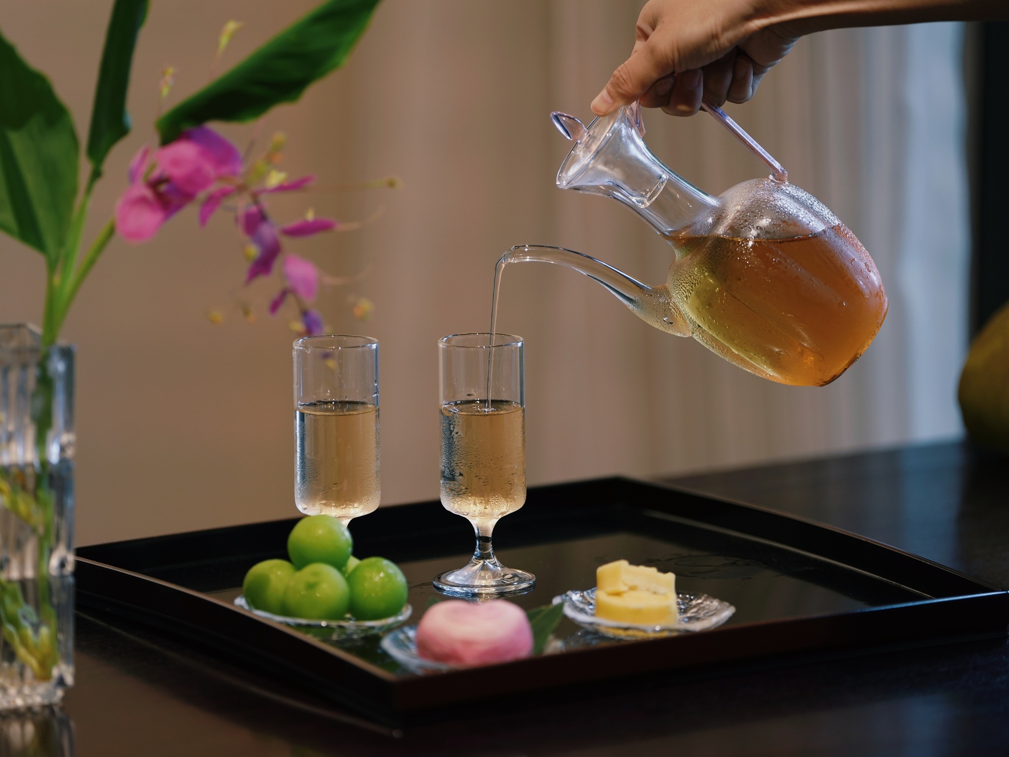 茶舍內，手將琥珀色茶飲注入杯，旁有鮮果、茶點，精緻誘人