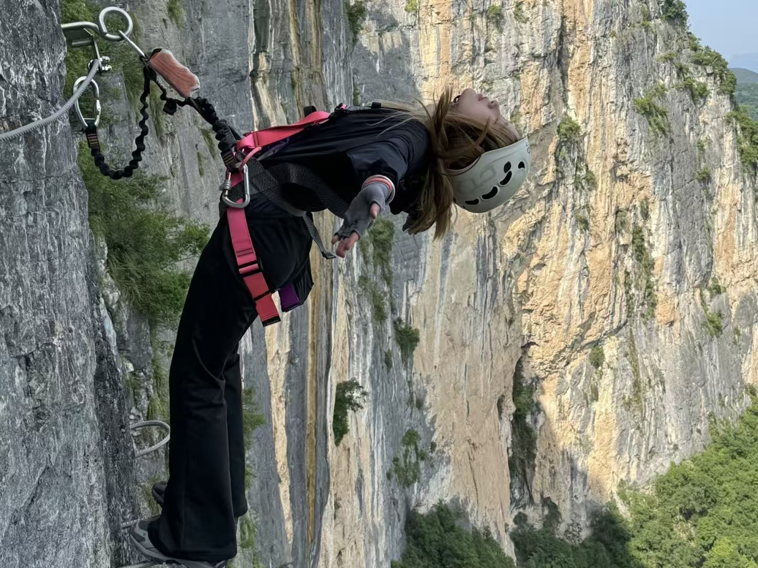 黑衣女子貼崖壁後仰，粉帶裝備襯著巖色肌理，髮絲紛飛，是懸崖邊與風共舞的酷感與自由