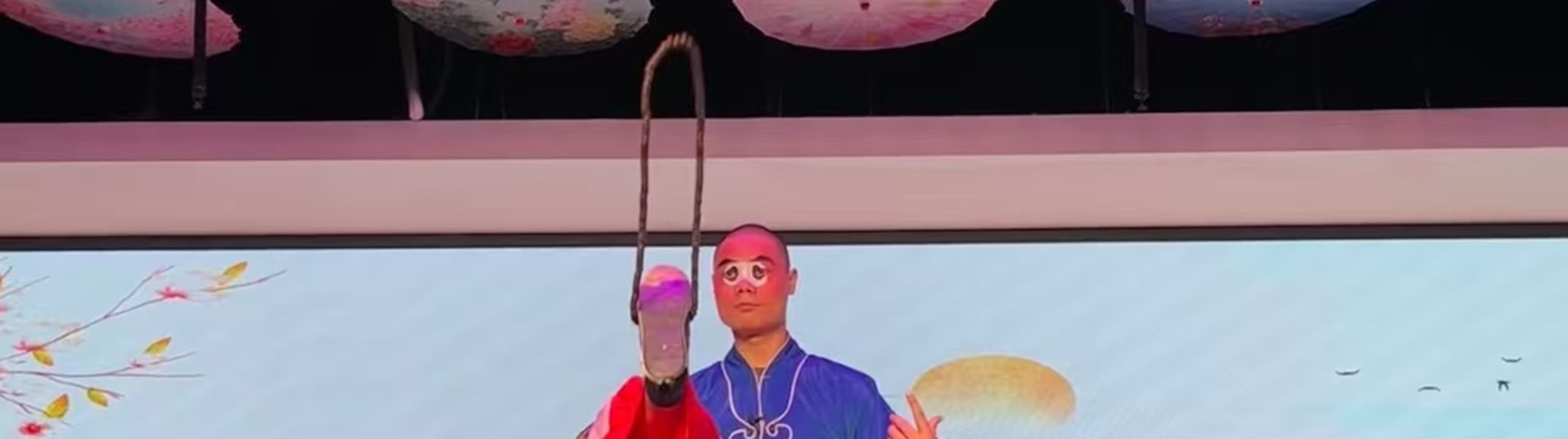 戲曲演員扮作光頭角色，著藍袍紅褲單腿高抬，旁有紅衣旦角佇立，背景是山水橋景，動作頗具張力