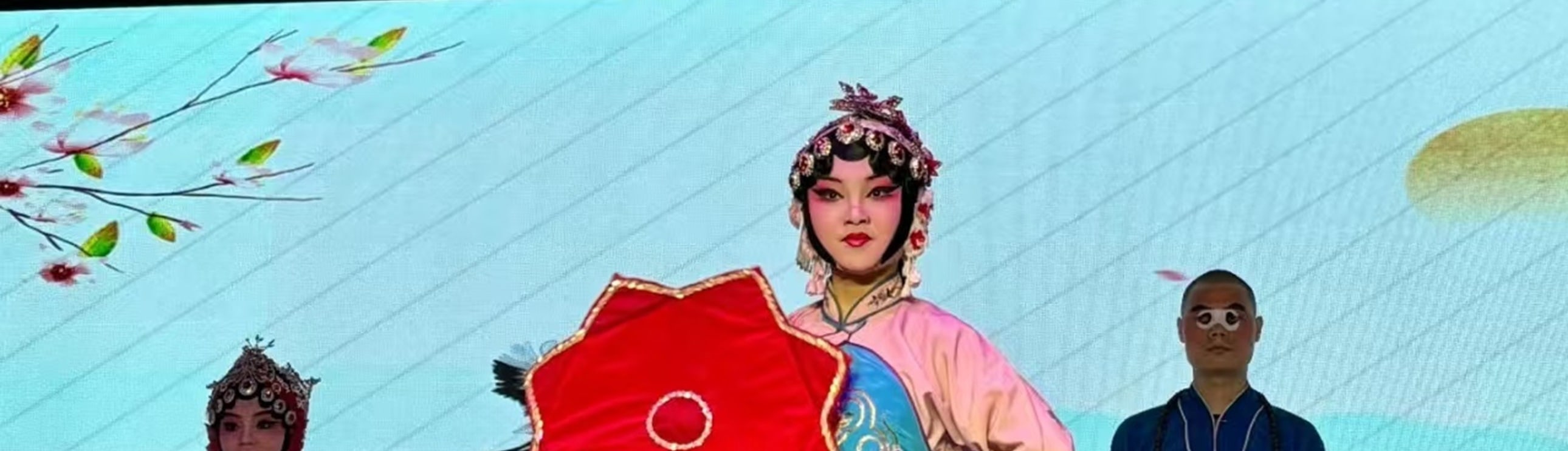 戲曲舞臺上，三位演員扮相鮮明：中間持紅花傘者衣飾繡花鳥，兩側分著紅衣、藍袍，背景配山水屏與傘飾，氛圍雅緻