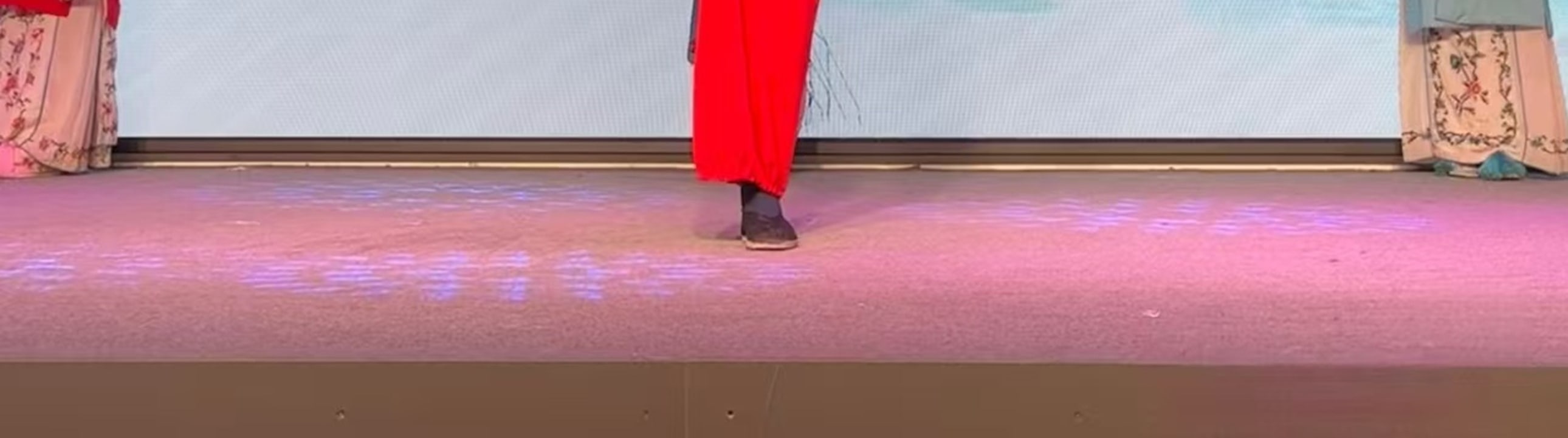 戲曲演員扮作光頭角色，著藍袍紅褲單腿高抬，旁有紅衣旦角佇立，背景是山水橋景，動作頗具張力