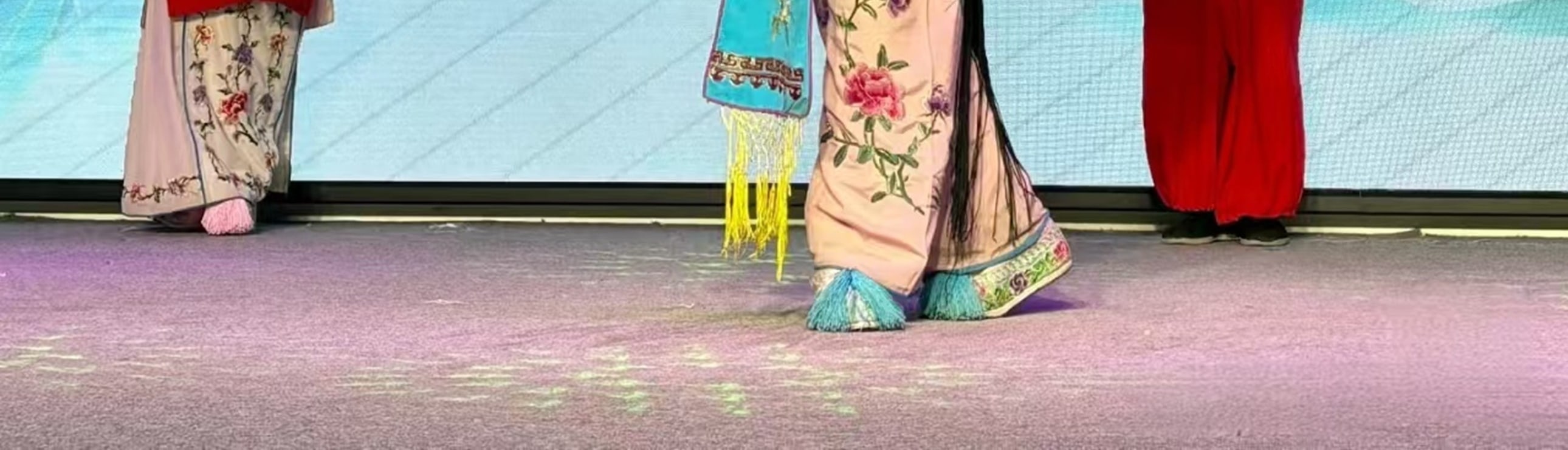 戲曲舞臺上，三位演員扮相鮮明：中間持紅花傘者衣飾繡花鳥，兩側分著紅衣、藍袍，背景配山水屏與傘飾，氛圍雅緻