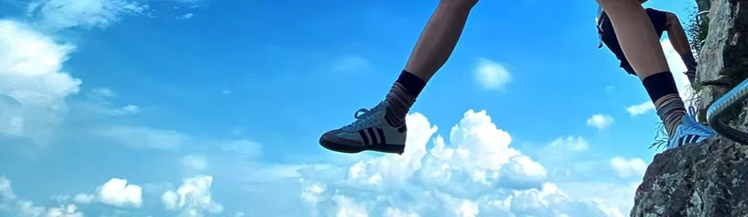 穿熒光綠衣的挑戰者展臂分腿，立於崖壁懸空設施上，藍天流雲作背景，安全裝備襯著舒展姿態，是高空挑戰裡的自由與酷感