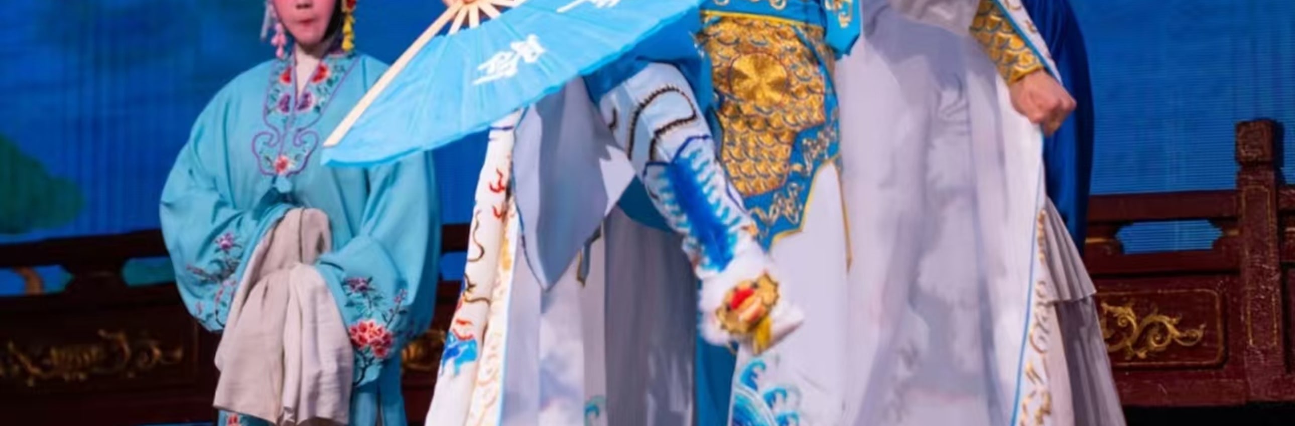 武生飾藍臉臉譜，著藍白金紋戲服，配翎子頭飾，單腿高抬展扇，旁有青衣旦角，盡顯戲曲武打的颯爽