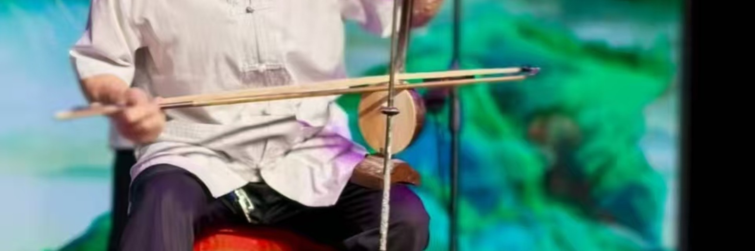 樂師持板胡演奏，著淺白衣褲，背景是青綠山水屏，神情投入，盡顯傳統樂器演奏的專注與韻味