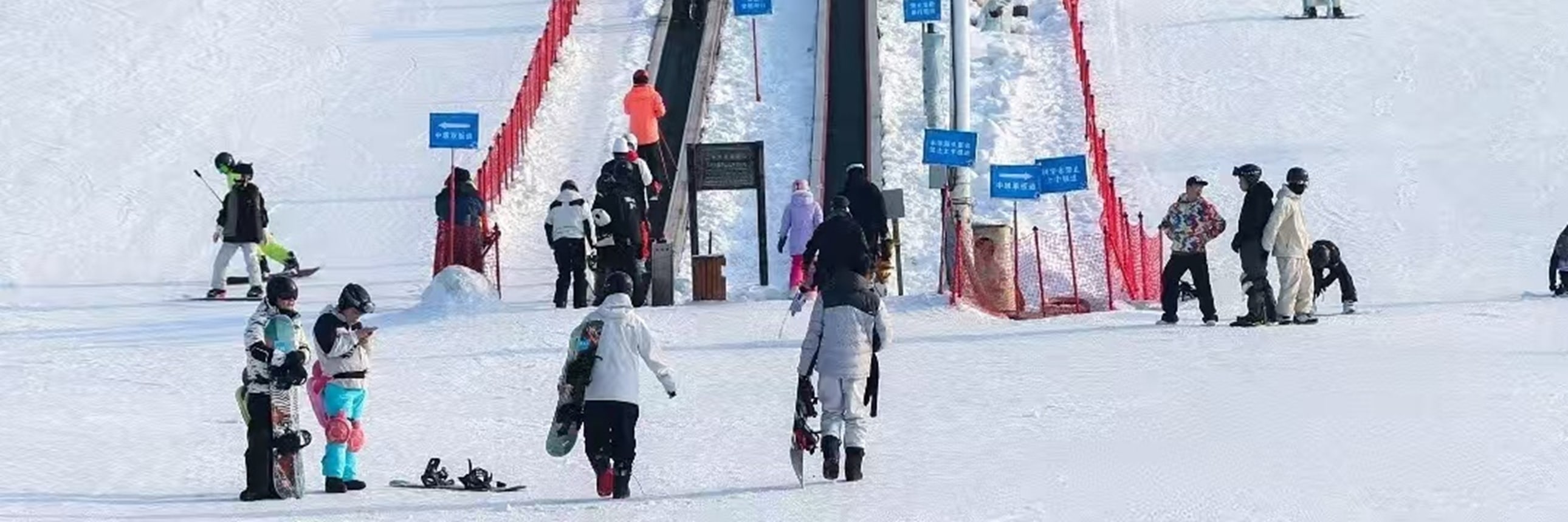 滑雪場雪道上人流攢動，中間魔毯傳送帶載著滑雪者向上，紅網圍欄分界，陽光鋪在雪面，熱鬧又有序