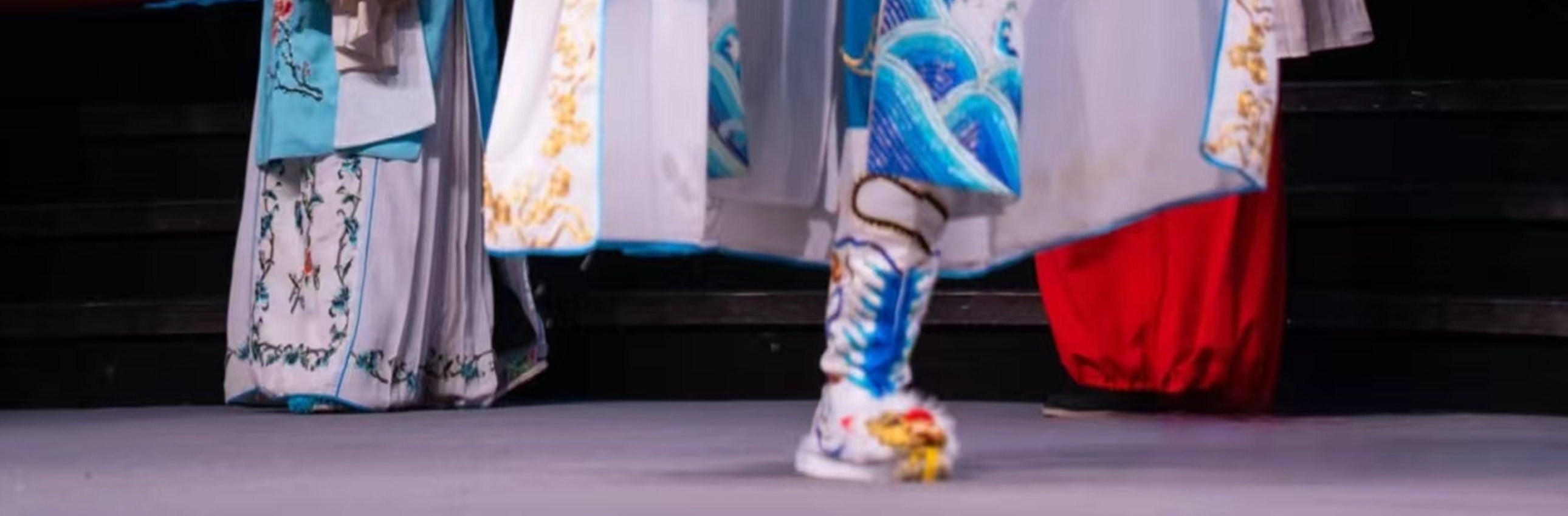 武生飾藍臉臉譜，著藍白金紋戲服，配翎子頭飾，單腿高抬展扇，旁有青衣旦角，盡顯戲曲武打的颯爽