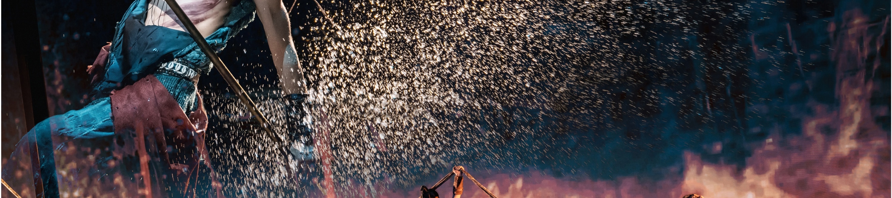 舞者手持長杆,在火花四濺的背景中展現出極具張力的動作,下方眾多舞者以繩索配合,整個場景充滿了動感與力量。 舞者手持長杆,在火花四濺的背景中展現出極具張力的動作,下方眾多舞者以繩索配合,整個場景充滿了動感與力量。