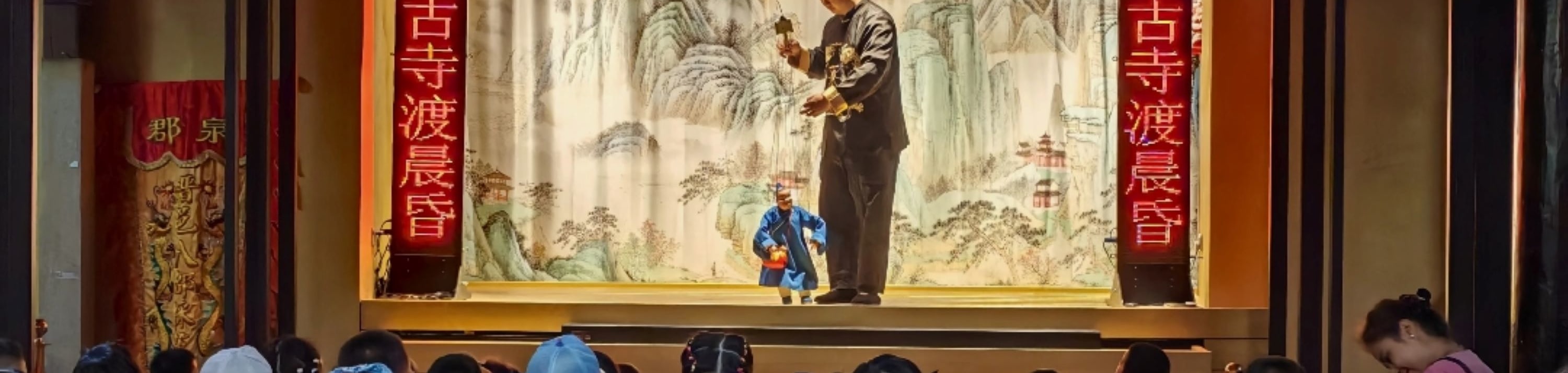 提線木偶戲演出現場，舞臺掛紅燈籠、配山水幕布，小觀眾們坐滿臺下，傳統民俗表演受孩童歡迎