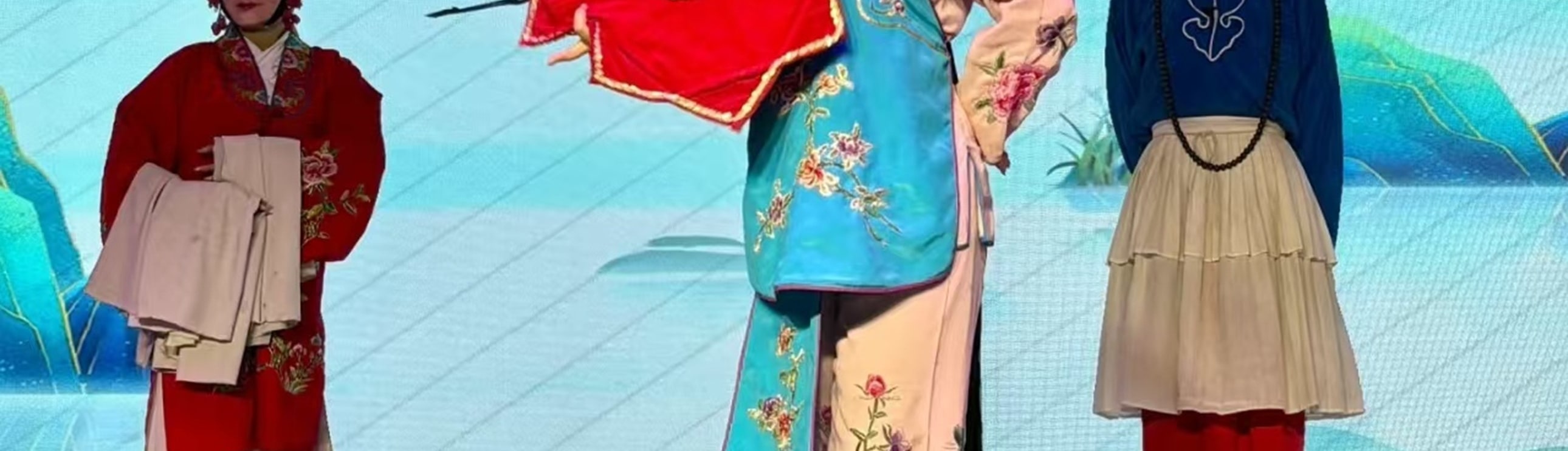 戲曲舞臺上，三位演員扮相鮮明：中間持紅花傘者衣飾繡花鳥，兩側分著紅衣、藍袍，背景配山水屏與傘飾，氛圍雅緻