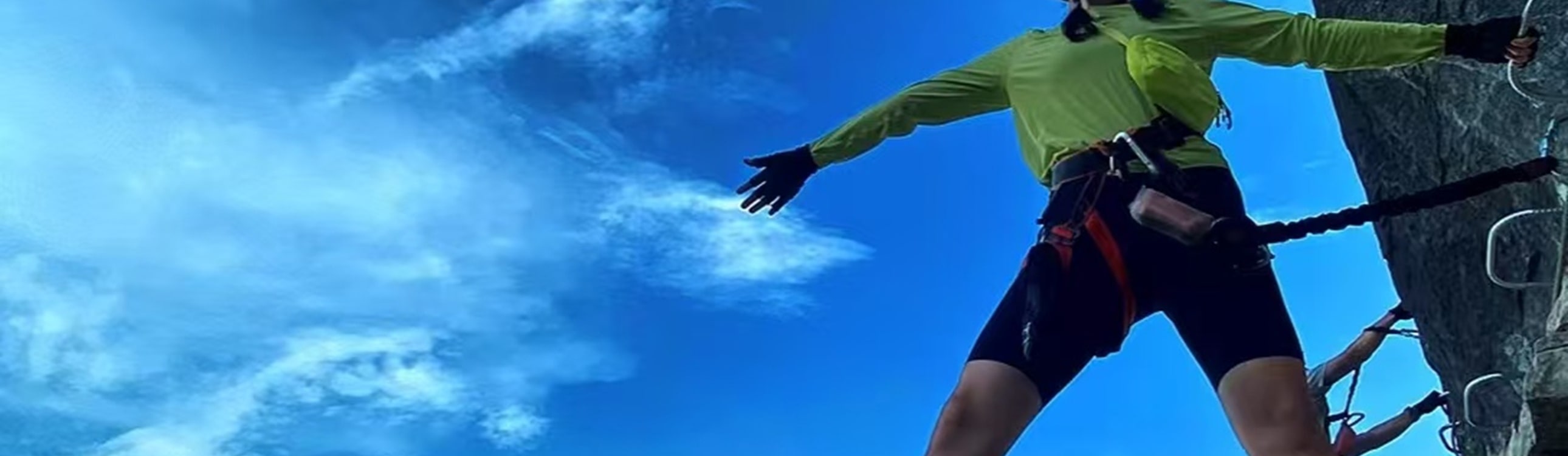 穿熒光綠衣的挑戰者展臂分腿，立於崖壁懸空設施上，藍天流雲作背景，安全裝備襯著舒展姿態，是高空挑戰裡的自由與酷感