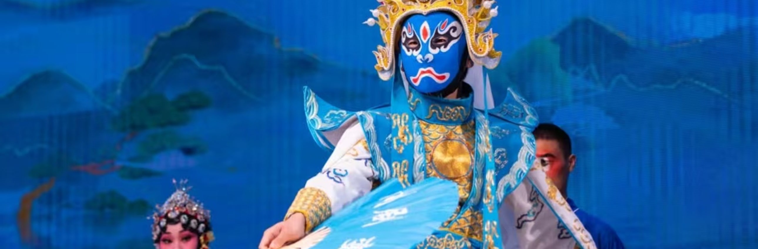 武生飾藍臉臉譜，著藍白金紋戲服，配翎子頭飾，單腿高抬展扇，旁有青衣旦角，盡顯戲曲武打的颯爽