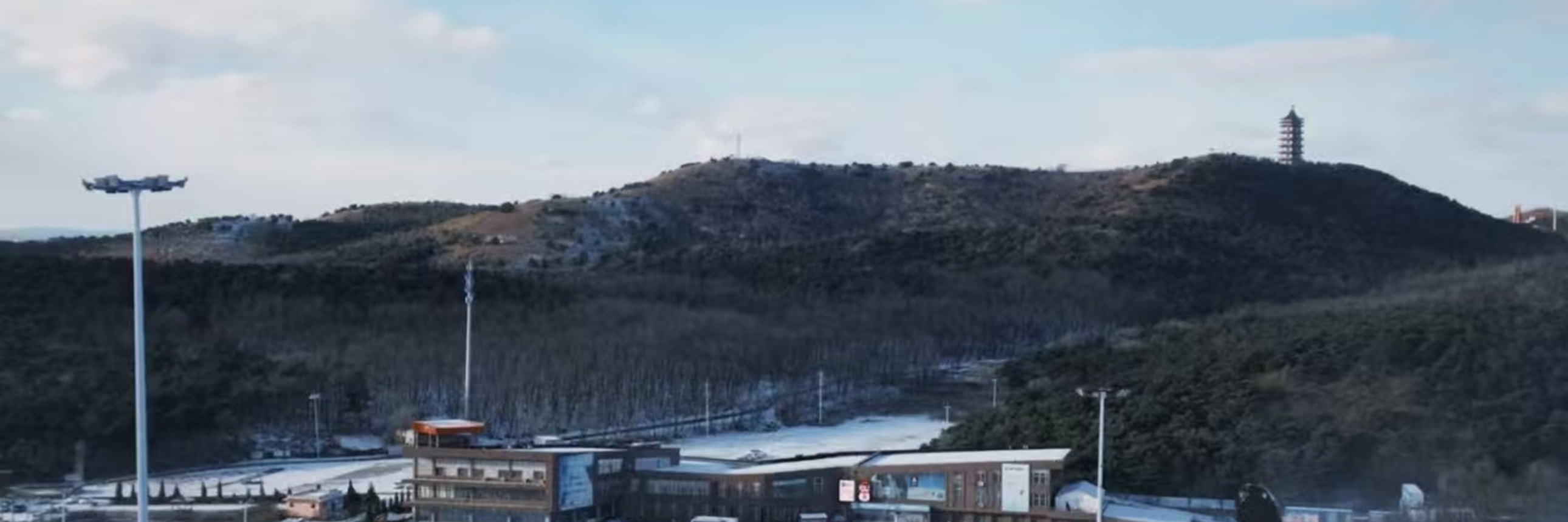 從雪坡頂俯瞰，雪場建築與人群錯落，遠山藏著塔影，近景雪具閒置，晴空襯得開闊雪場既熱鬧又舒展