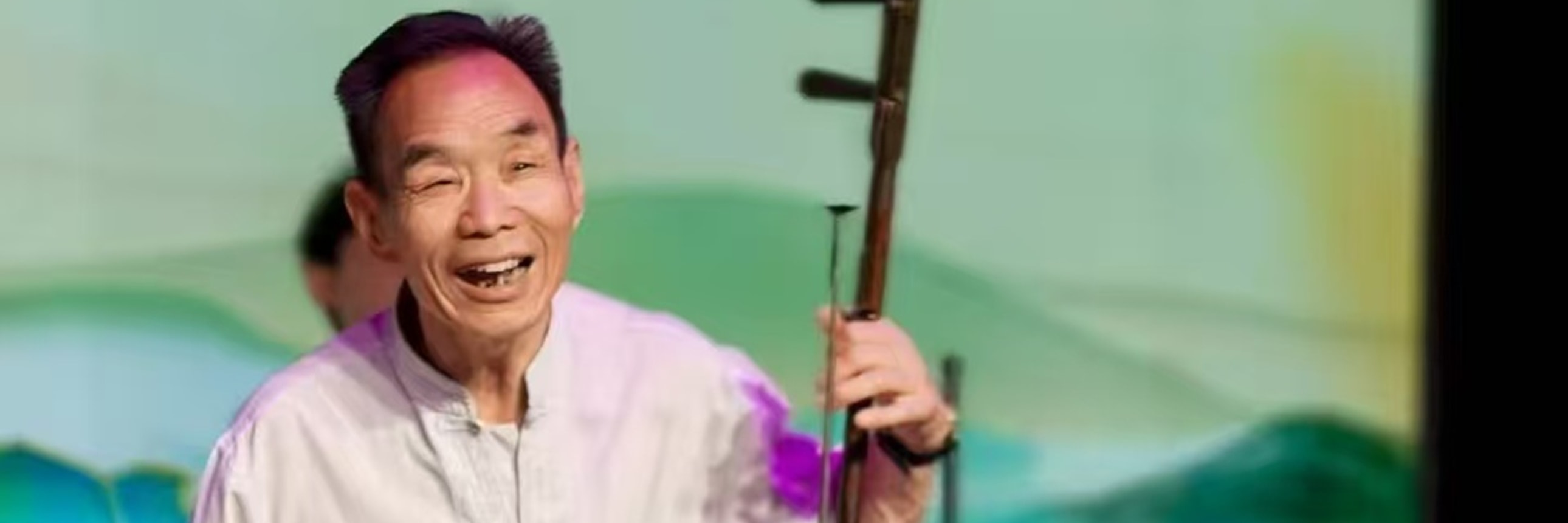 樂師持板胡演奏，著淺白衣褲，背景是青綠山水屏，神情投入，盡顯傳統樂器演奏的專注與韻味