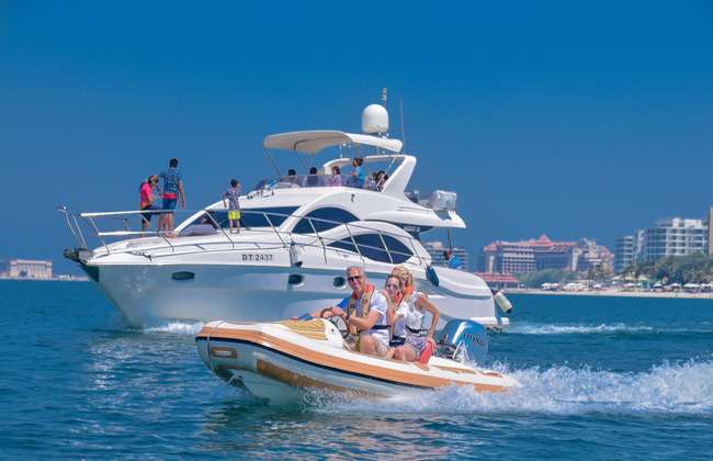 Two passengers pilot a compact speedboat on the water with a yacht and the Dubai shoreline behind them.