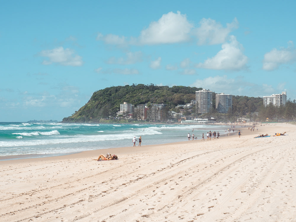 People lying on the sand of Burleigh Heads, with the city’s buildings seen from afar